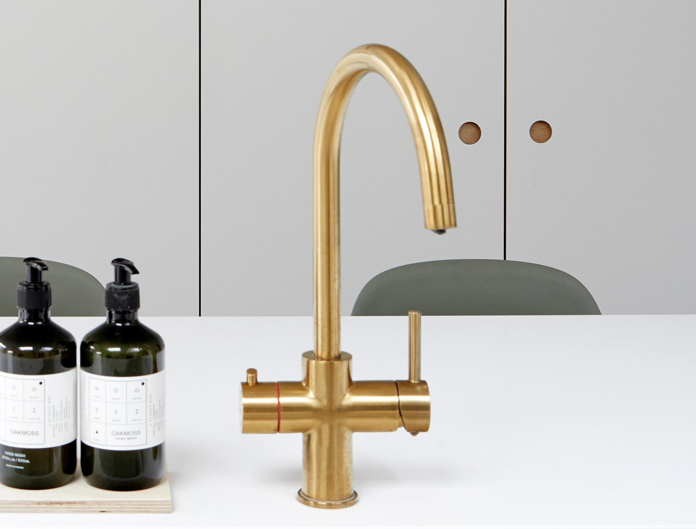 A modern gold kitchen faucet with a curved spout stands on a white countertop. Two pump bottles are placed to the left, and gray cabinets with round handles are in the background.