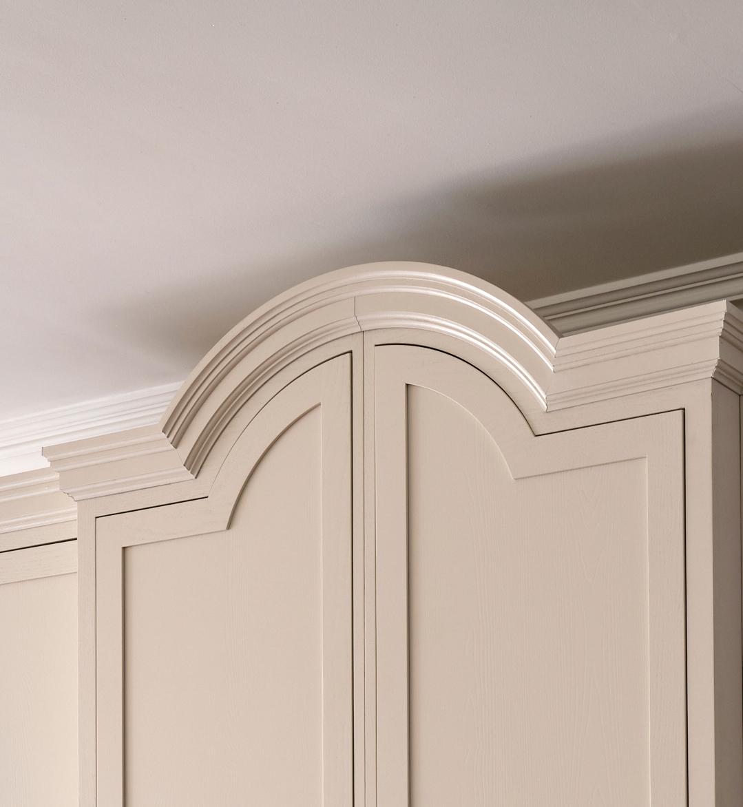 The image shows the upper section of a cream-colored cabinet with decorative crown molding and an arched, raised panel design near the ceiling. The wall and ceiling are painted in light, neutral tones.
