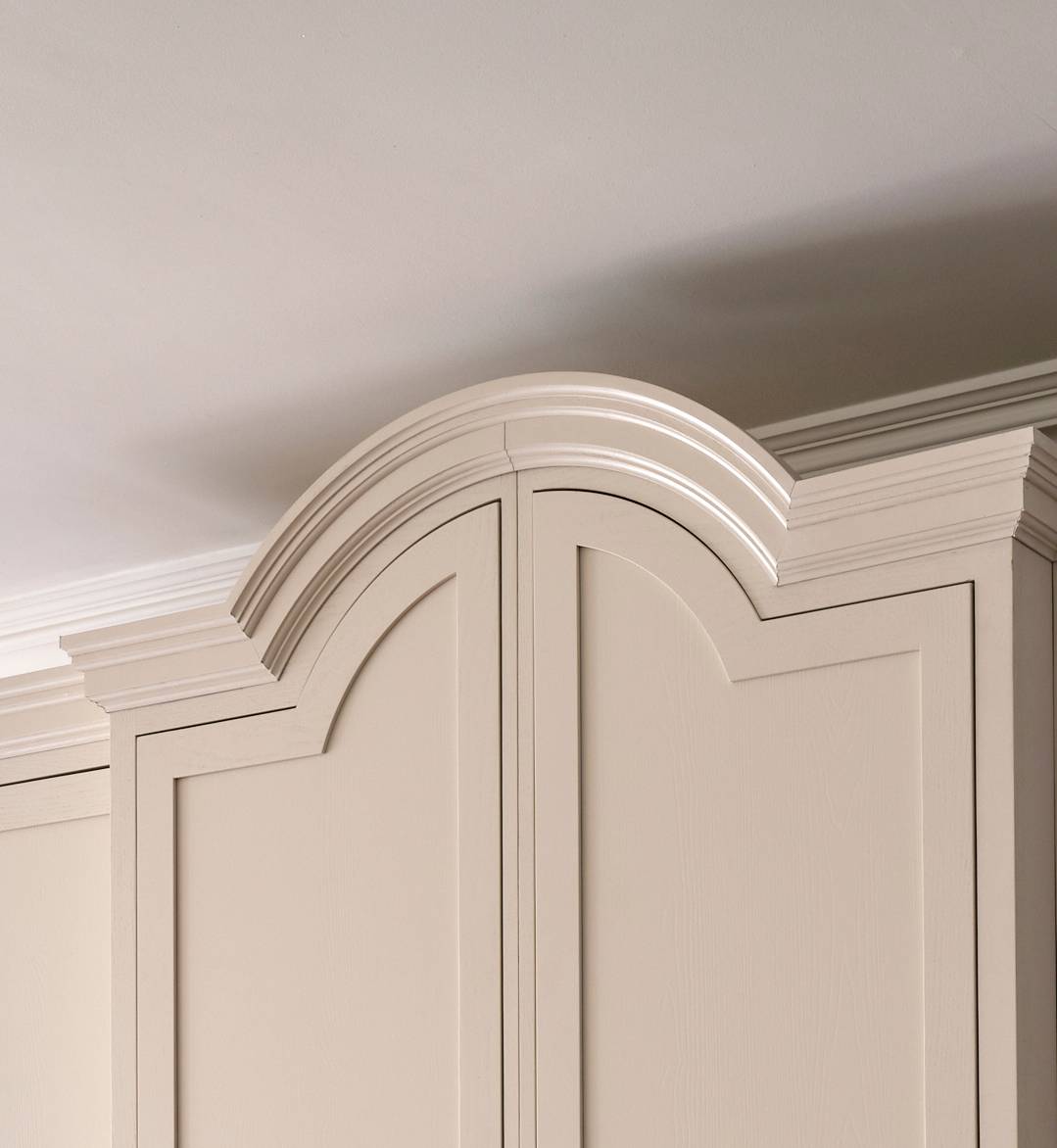 The image shows the upper section of a cream-colored cabinet with decorative crown molding and an arched, raised panel design near the ceiling. The wall and ceiling are painted in light, neutral tones.