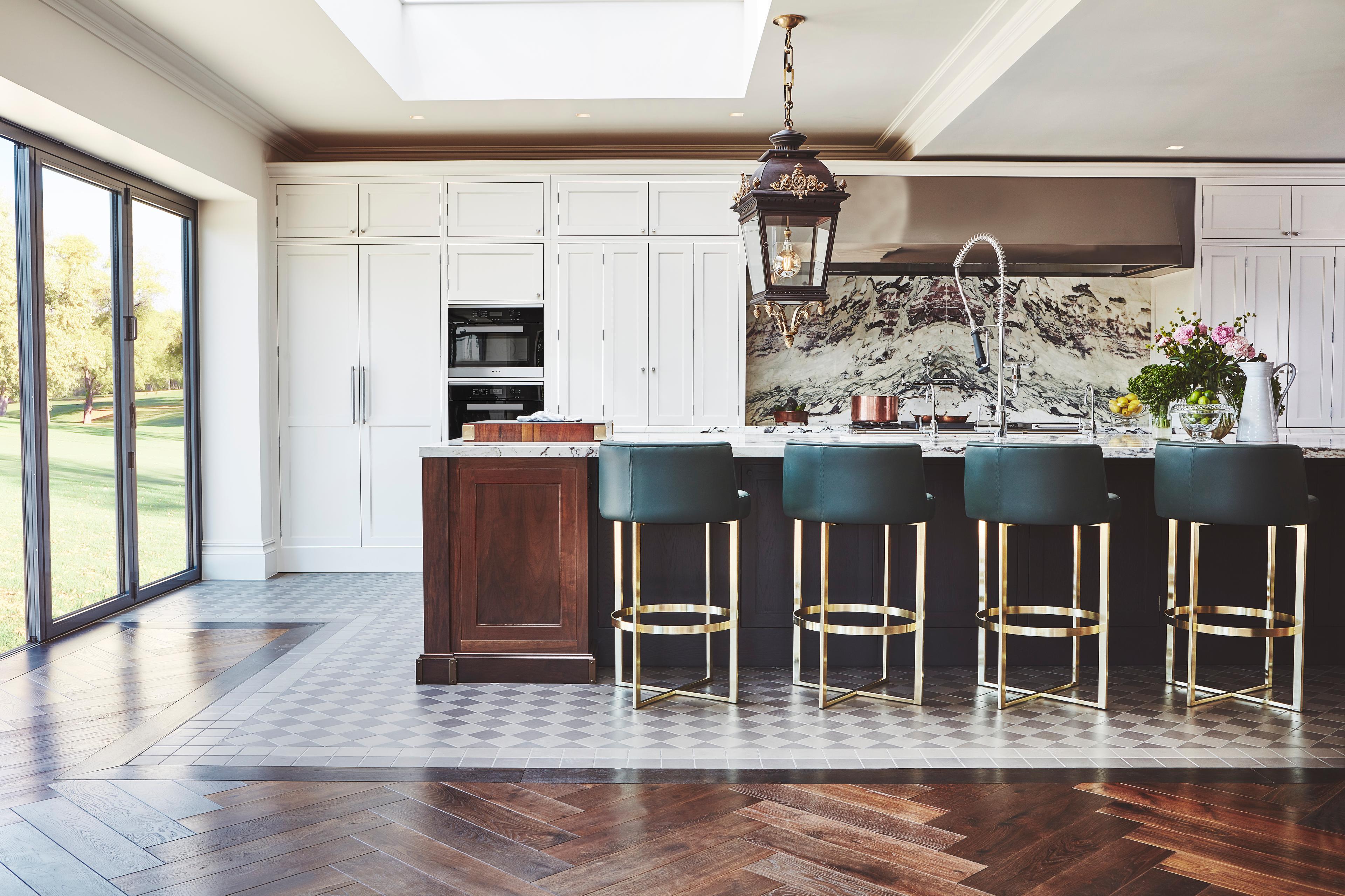 a luxurious, spacious kitchen features a large dark island lined with four teal upholstered bar stools on gold legs, an ornate hanging lantern, a dramatic marble backsplash, and large sliding glass doors opening onto a bright lawn.