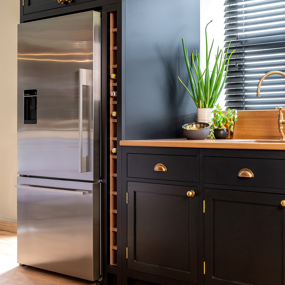 A modern kitchen with a stainless steel refrigerator, vertical wine rack, black cabinets with brass handles, a wooden countertop, houseplants, and a brass faucet by a window with black blinds.