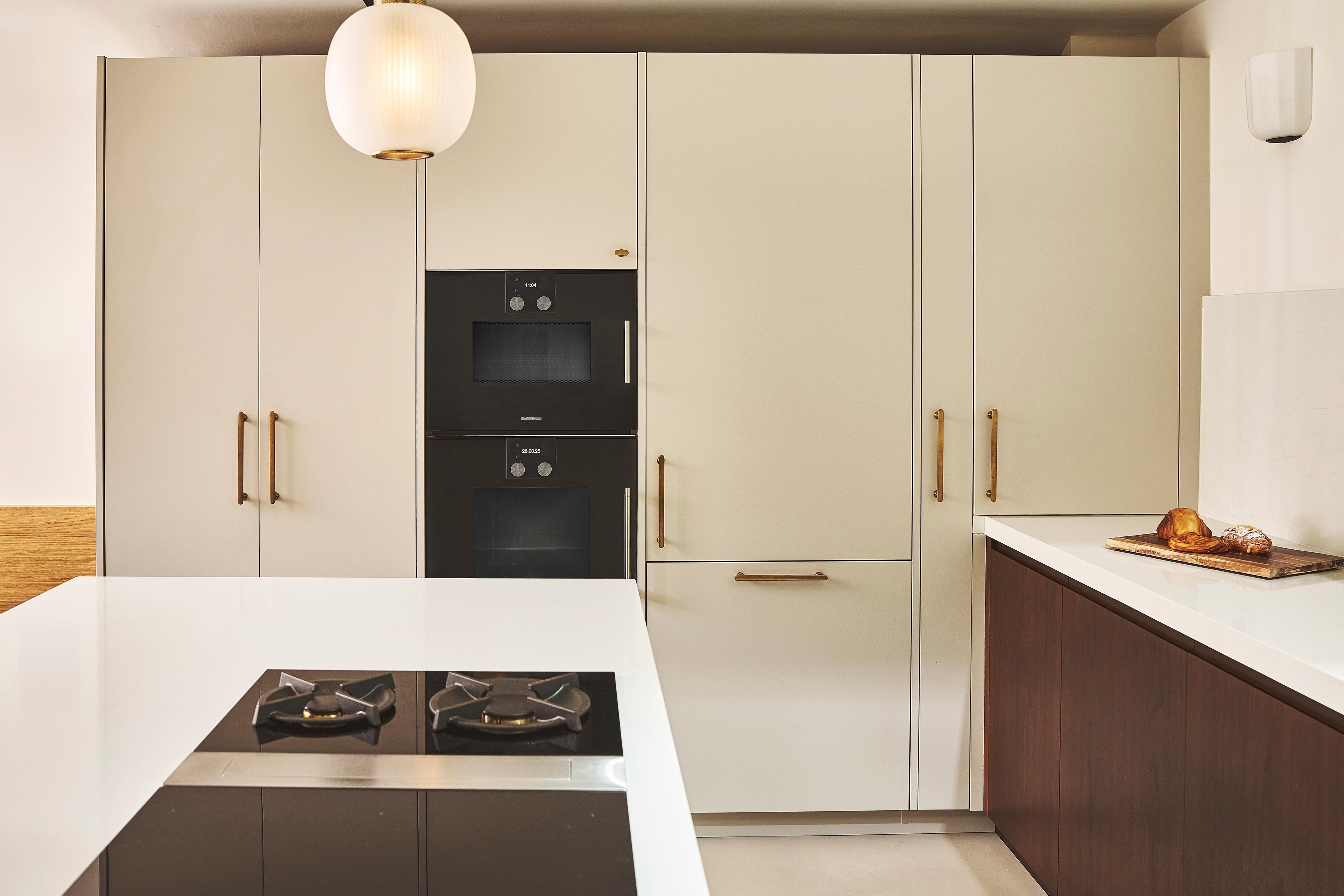 Modern kitchen with white cabinets, built-in black oven, gold handles, a gas stove on a white island, and a countertop with a cutting board and bread. A round pendant light hangs from the ceiling