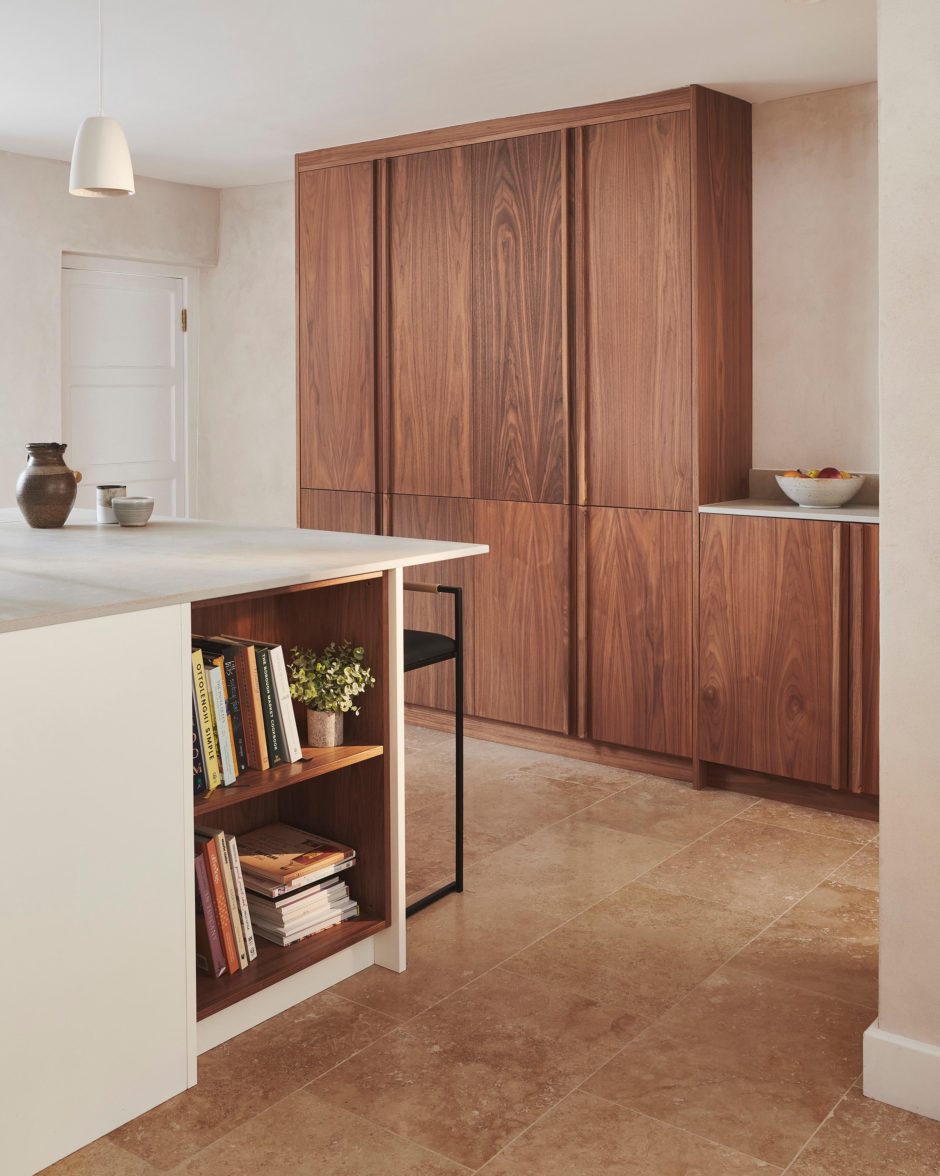 Modern kitchen with a minimalist design. Features wooden cabinets, a large island with a light countertop, and a few decorative items. Open shelving on island holds books. Beige stone tile flooring enhances the warm, natural aesthetic.
