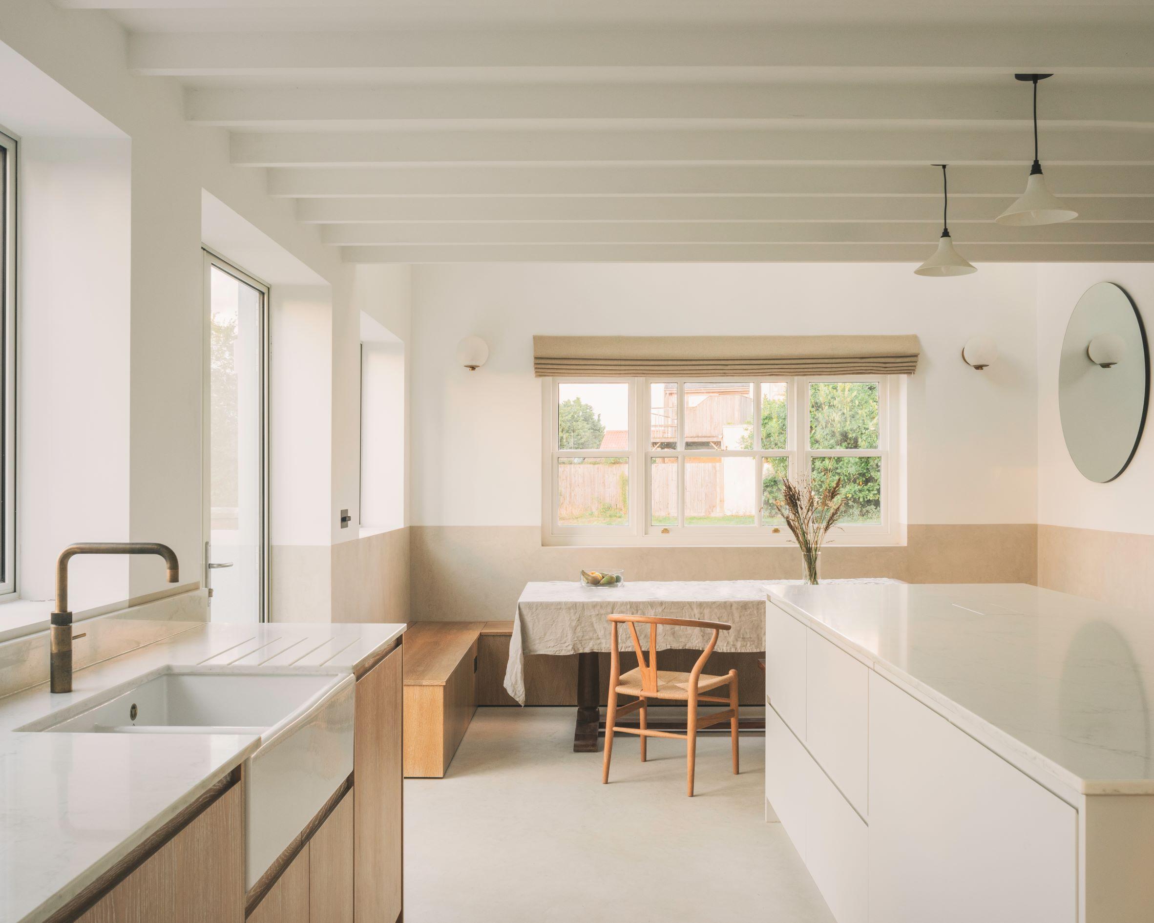Minimalist kitchen with white walls, large windows, a central island, built-in sink, and wooden accents. A dining table with a single chair sits by the window; natural light fills the space.