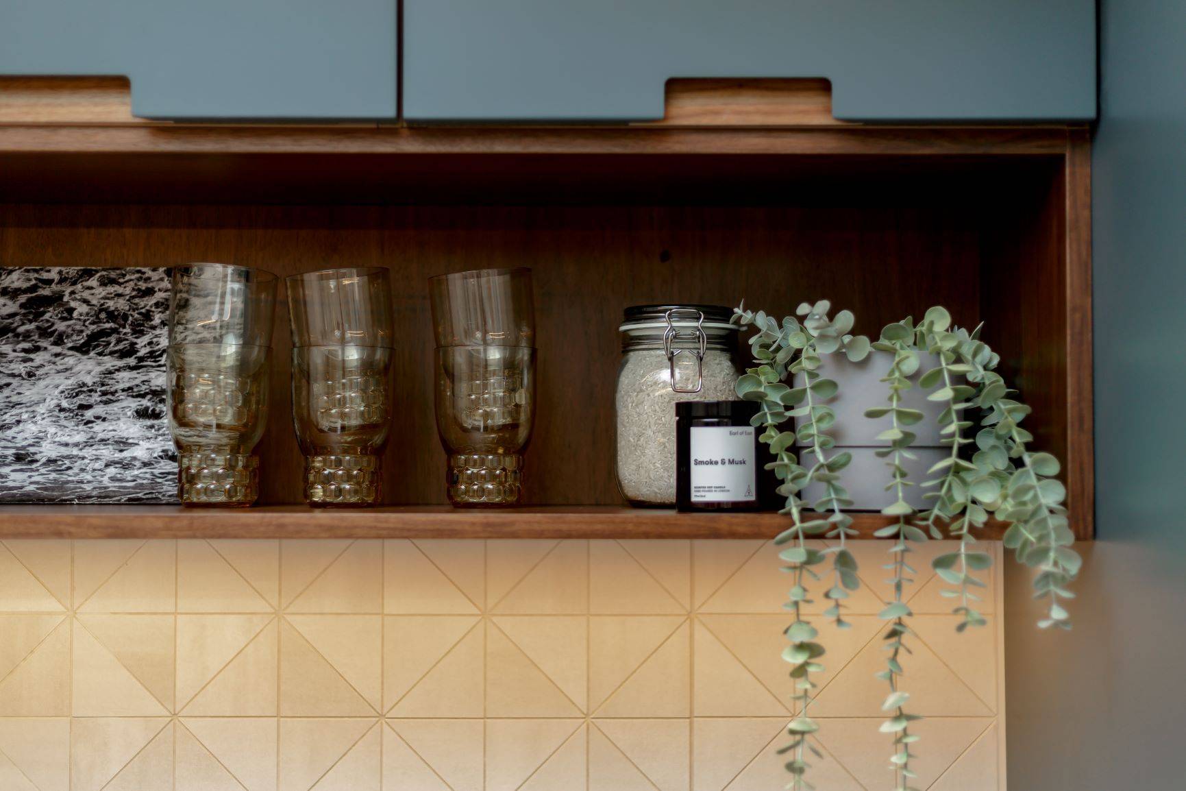 A wooden kitchen shelf displays translucent glasses and a candle next to a potted plant with cascading green leaves. The backdrop features geometric beige tiles.