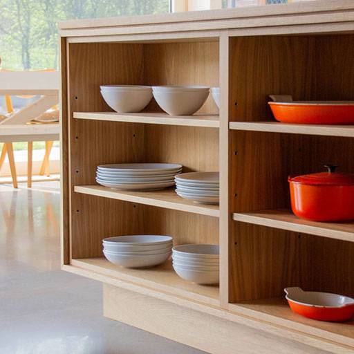 A wooden kitchen shelf displays white bowls and plates neatly stacked, along with bright orange pots and dishes. Sunlight streams in from a window, and a table with chairs is visible in the background.
