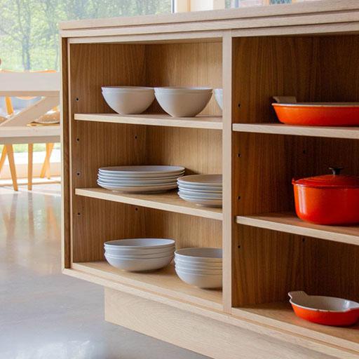 A wooden kitchen shelf displays white bowls and plates neatly stacked, along with bright orange pots and dishes. Sunlight streams in from a window, and a table with chairs is visible in the background.