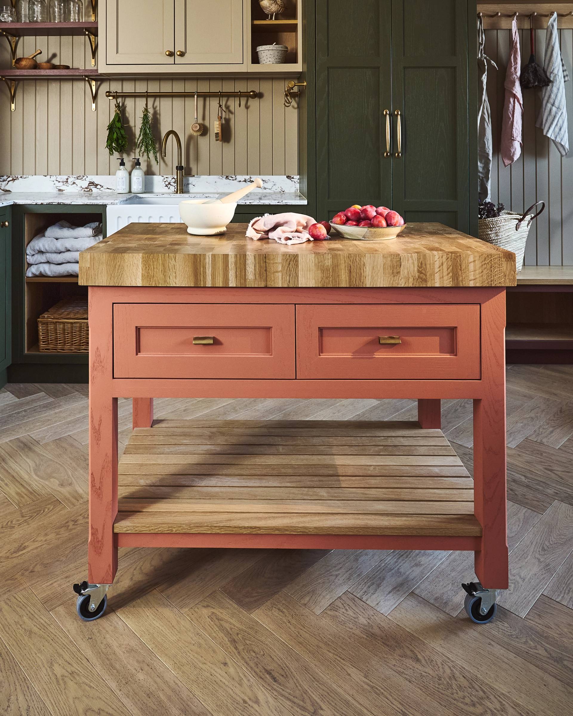 A small pink island unit on caster wheels with two drawers and a thick wooden worktop. On top there is a mixing bowl and a bowl of apples and a towel. In the background there is dark green cabinetry.
