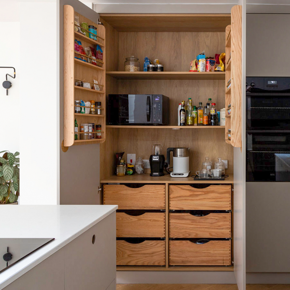 Open pantry with wooden shelves and drawers, stocked with jars, cans, spices, and kitchen appliances like a microwave, coffee machine, and blender. Pantry doors have spice racks; the modern kitchen is visible nearby.