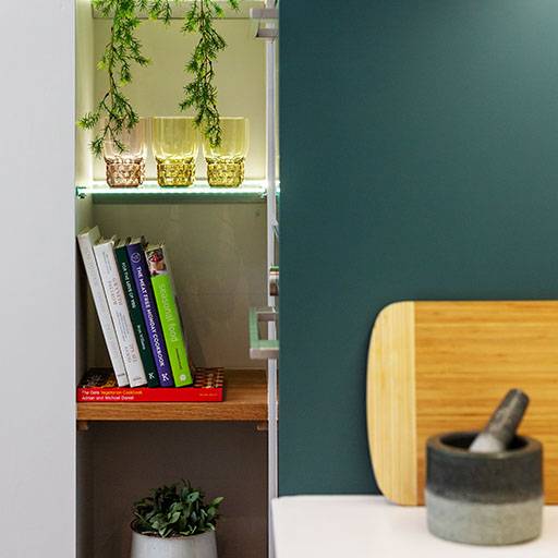 A small kitchen shelf holds glass cups, trailing greenery, and a row of cookbooks. Nearby are a wooden cutting board, a stone mortar and pestle, and a potted plant on a lower shelf.
