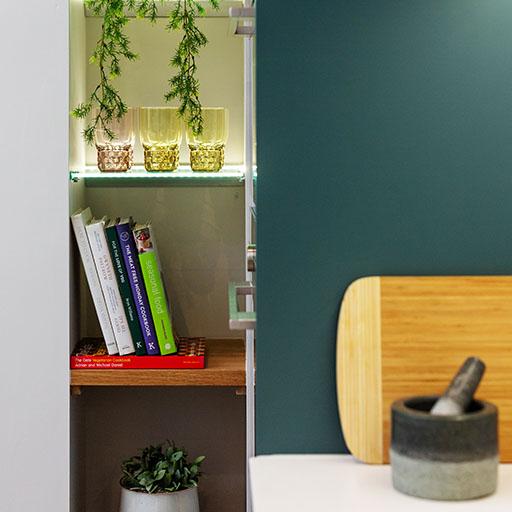 A small kitchen shelf holds glass cups, trailing greenery, and a row of cookbooks. Nearby are a wooden cutting board, a stone mortar and pestle, and a potted plant on a lower shelf.