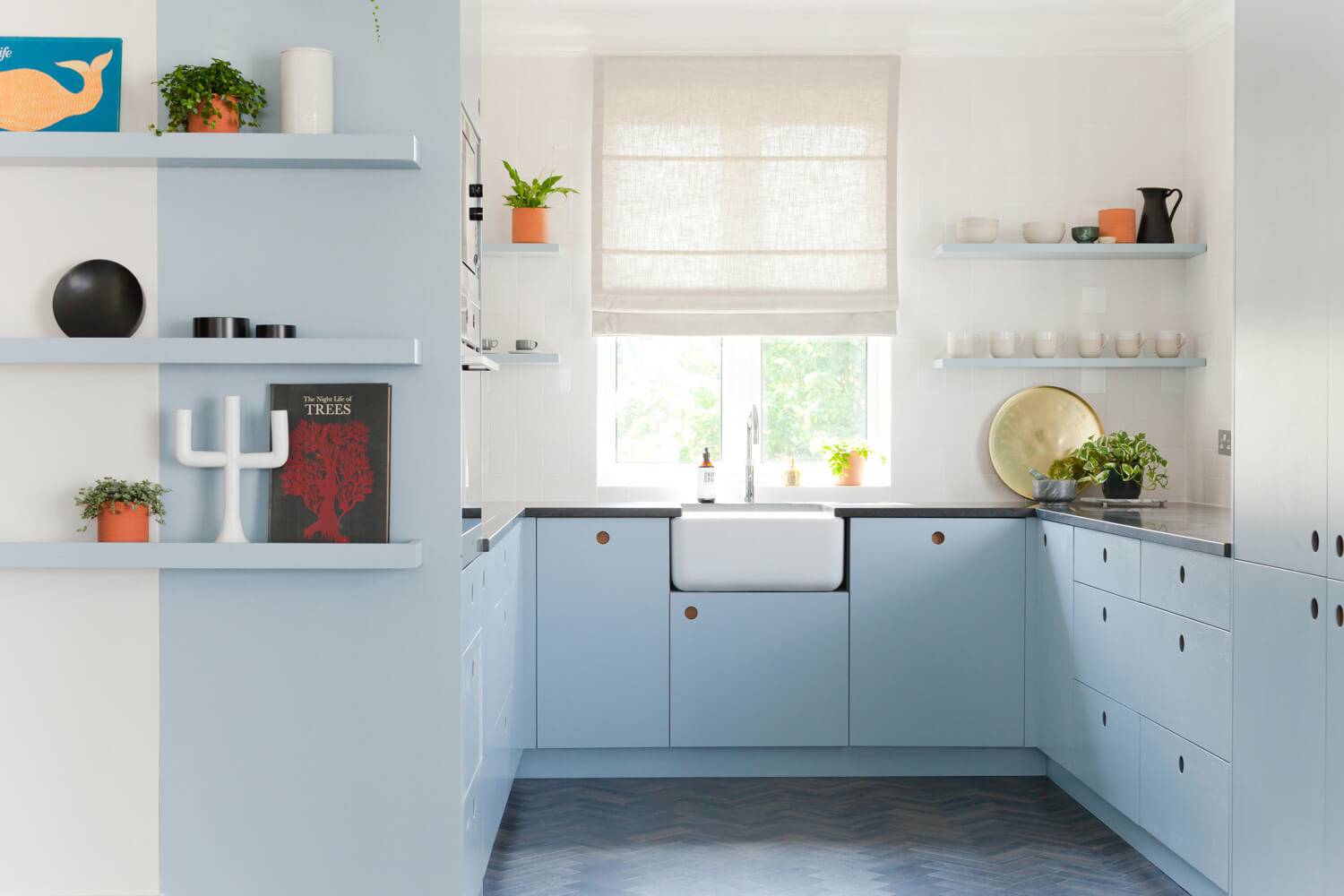 A modern kitchen with pale blue cabinets, a large farmhouse sink, and a window with a beige blind. Open shelves display plants, books, and decor. The floor features a dark herringbone pattern.