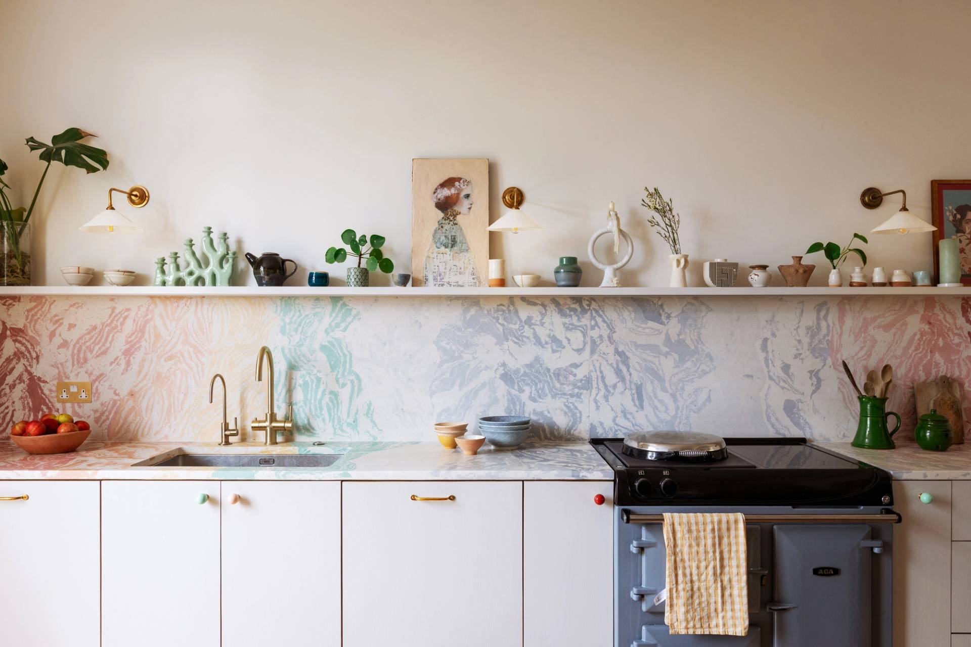 A colorful kitchen features a marble backsplash and countertops. It includes a sink with a gold faucet, a blue stove, various ceramic items, plants, and artwork on a shelf. A striped towel hangs on the stove handle.