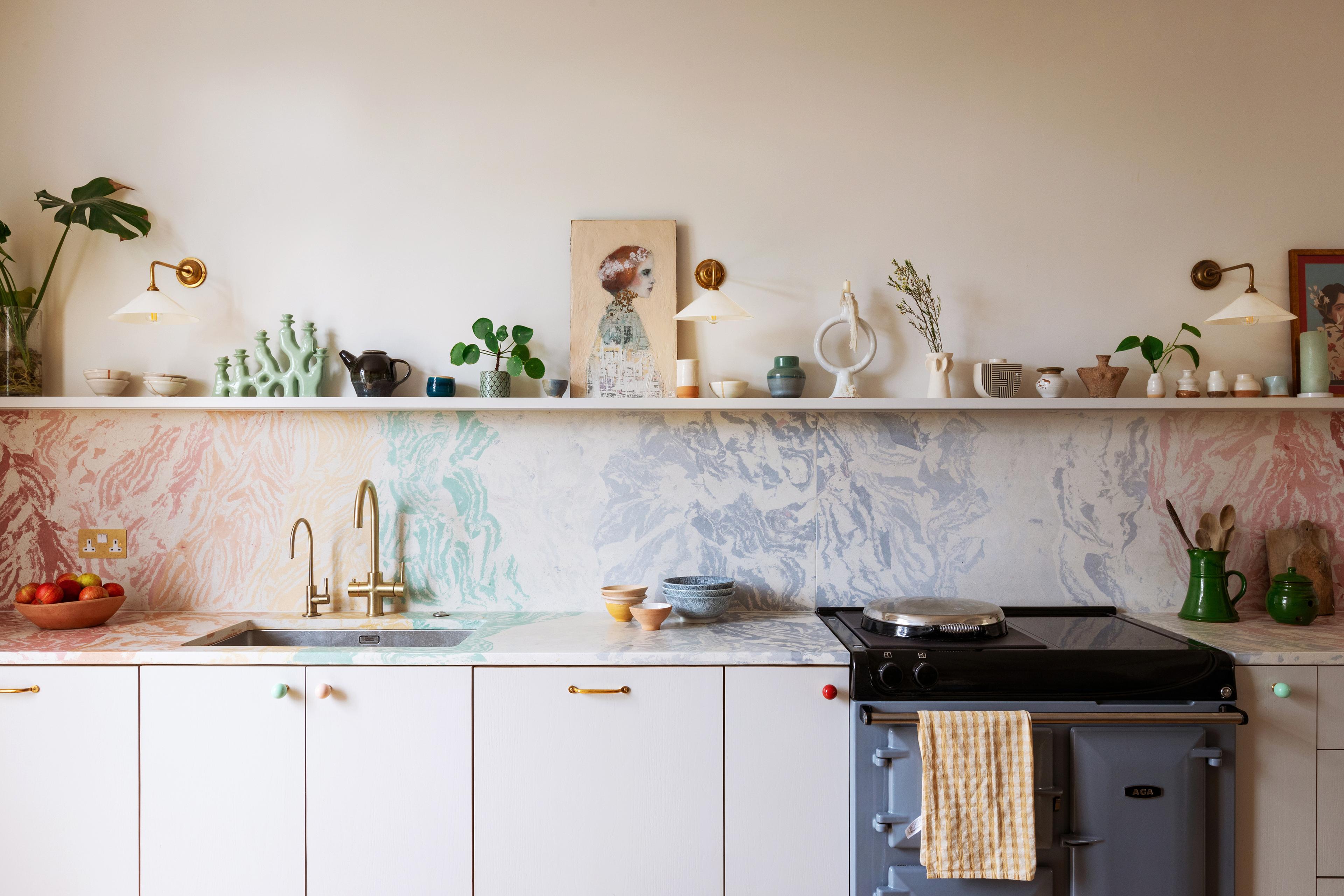 A colorful kitchen features a marble backsplash and countertops. It includes a sink with a gold faucet, a blue stove, various ceramic items, plants, and artwork on a shelf. A striped towel hangs on the stove handle.