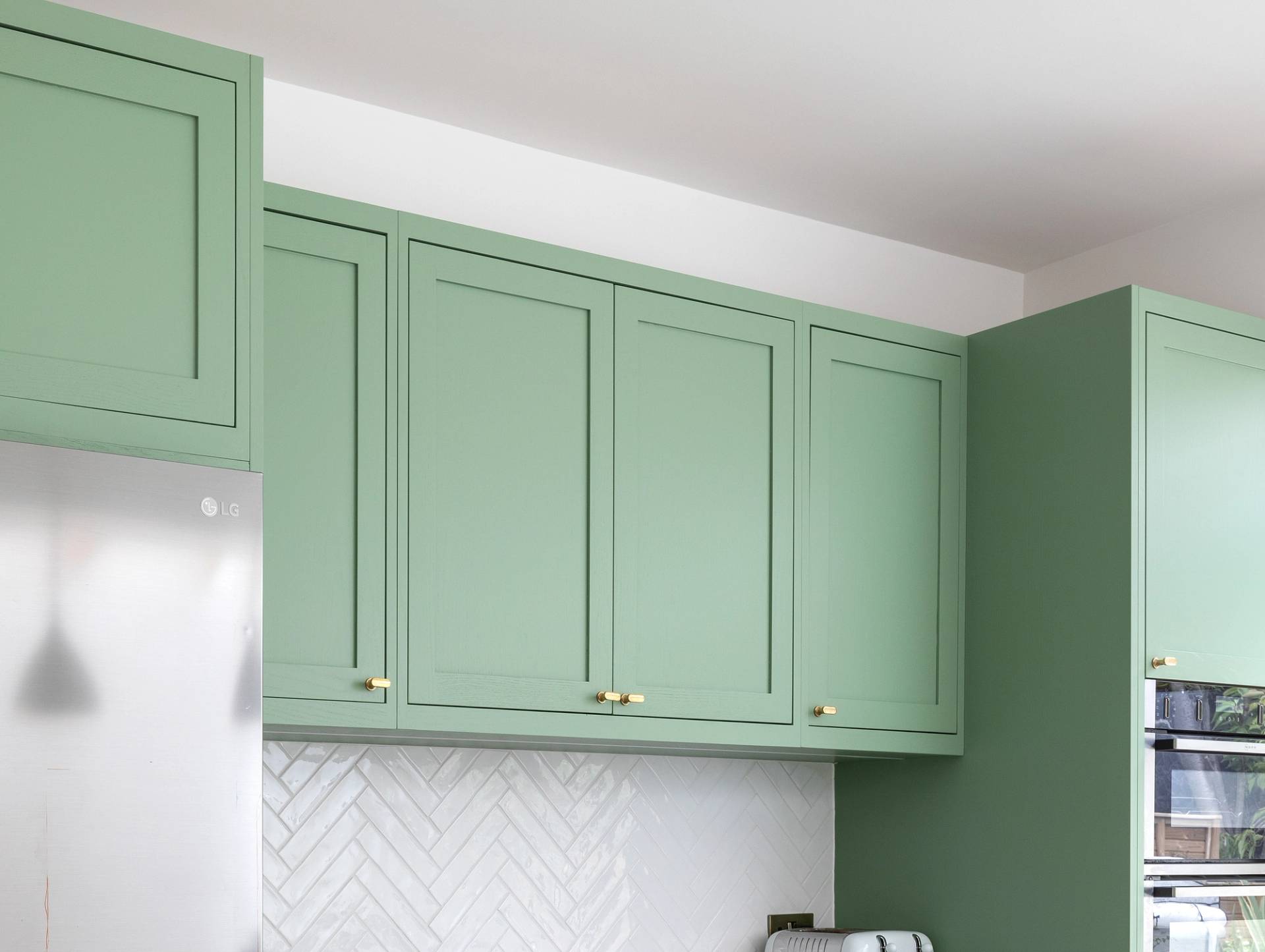 Green kitchen cabinets with gold handles are mounted above a white countertop and a chevron-patterned white tile backsplash. A sliver of a stainless steel refrigerator is visible on the left.
