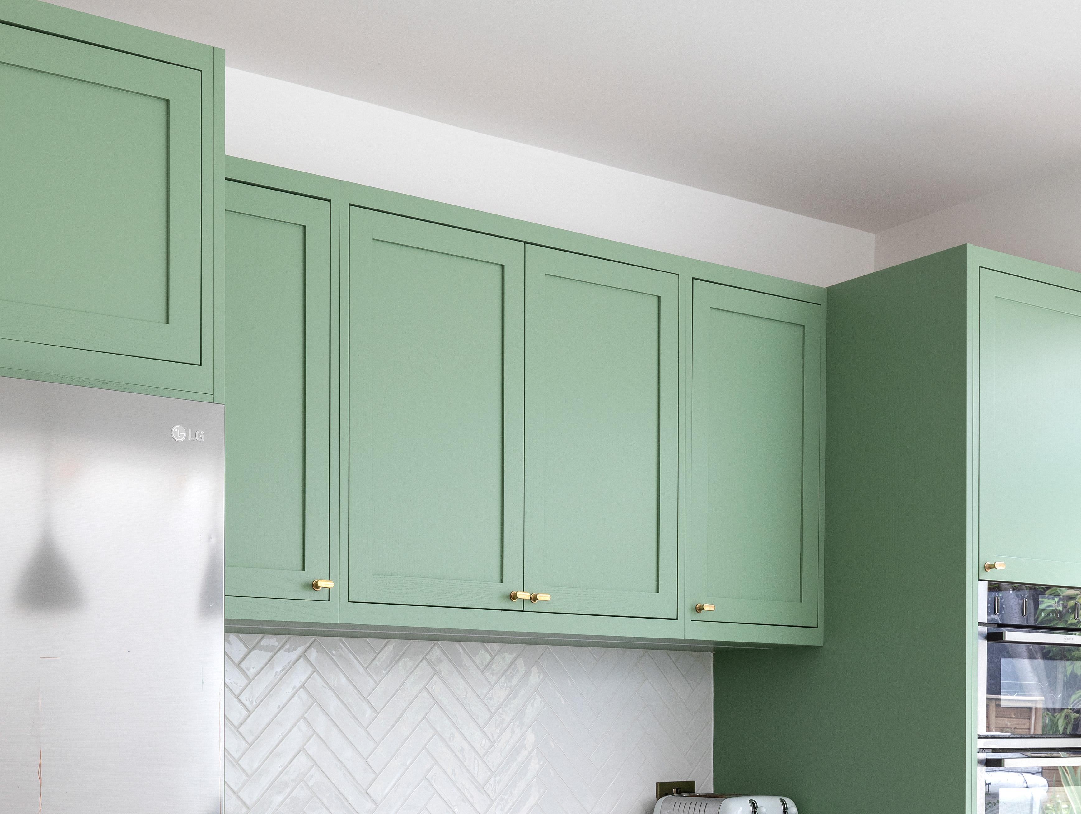 Green kitchen cabinets with gold handles are mounted above a white countertop and a chevron-patterned white tile backsplash. A sliver of a stainless steel refrigerator is visible on the left.