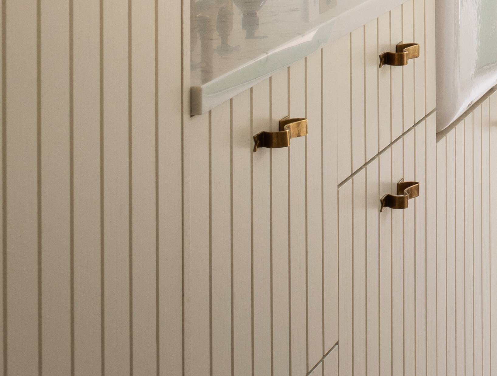 Close-up of cream-colored cabinets with vertical grooves and gold rectangular handles, topped with a light-colored countertop, next to a frosted glass window.