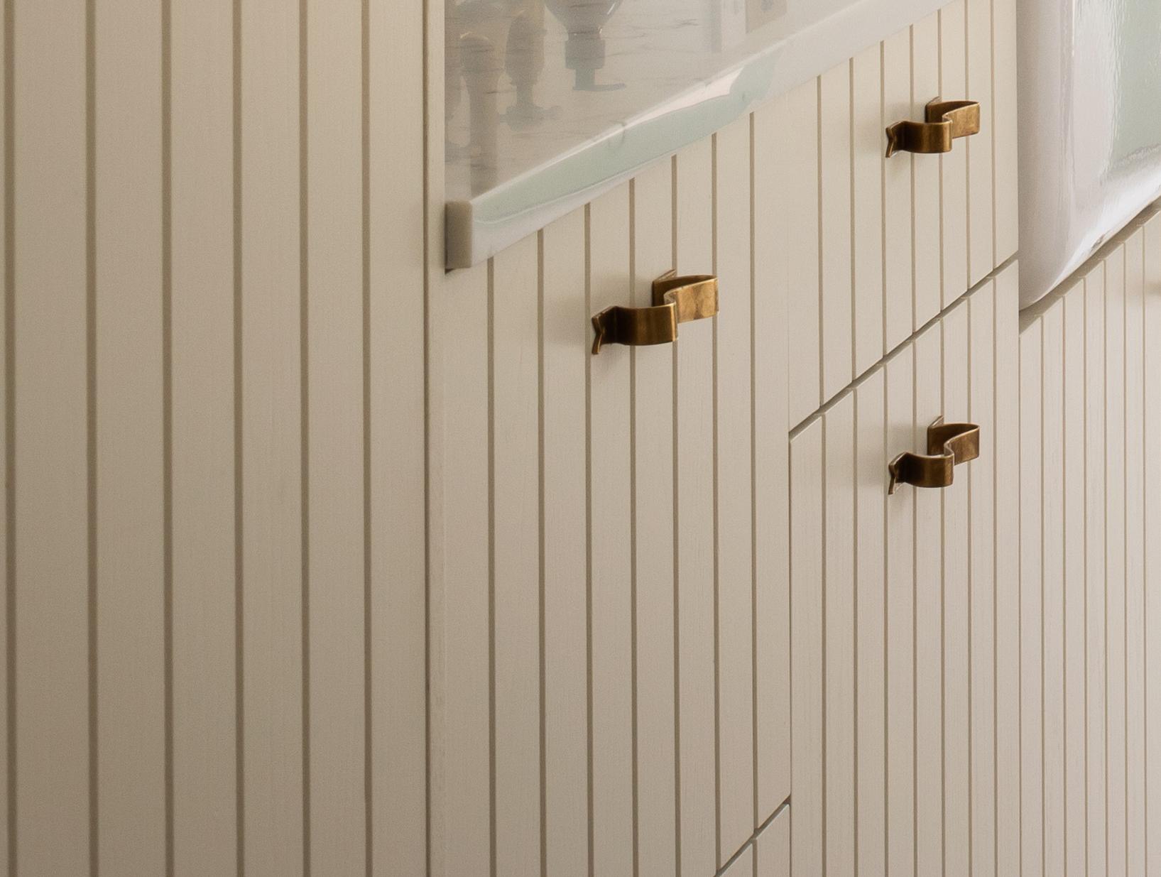Close-up of cream-colored cabinets with vertical grooves and gold rectangular handles, topped with a light-colored countertop, next to a frosted glass window.