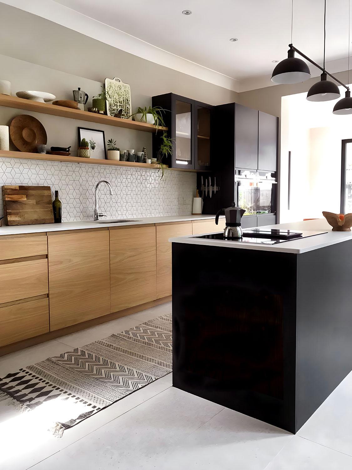 A modern kitchen with light wood cabinets, a black island, white tiled backsplash, open shelves with decor, a patterned rug, and pendant lights. Natural light fills the space through large windows.
