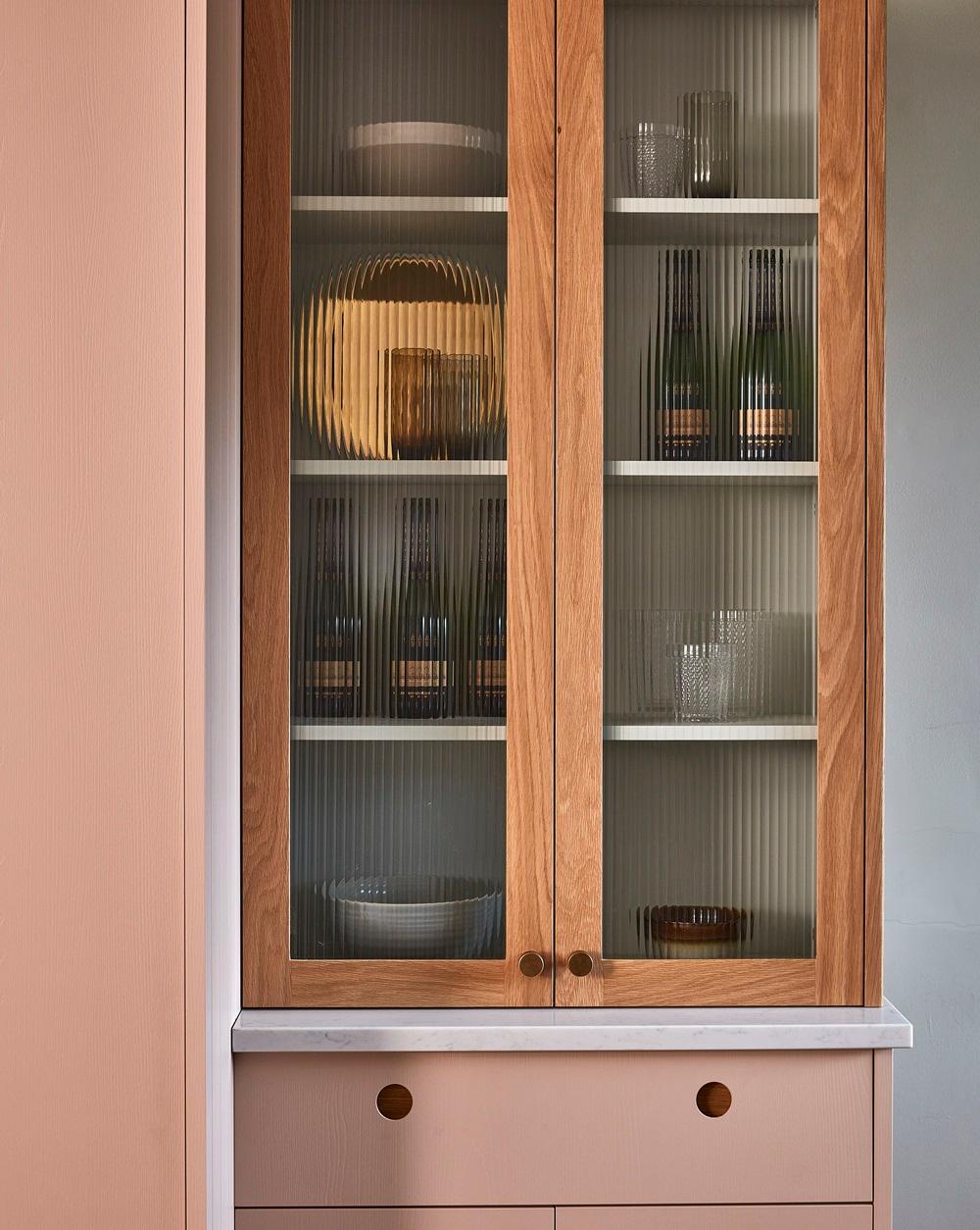 A wooden wall-mounted display cabinet with reeded glass doors resting above a pink base cabinet with round recessed handles and a marble countertop.