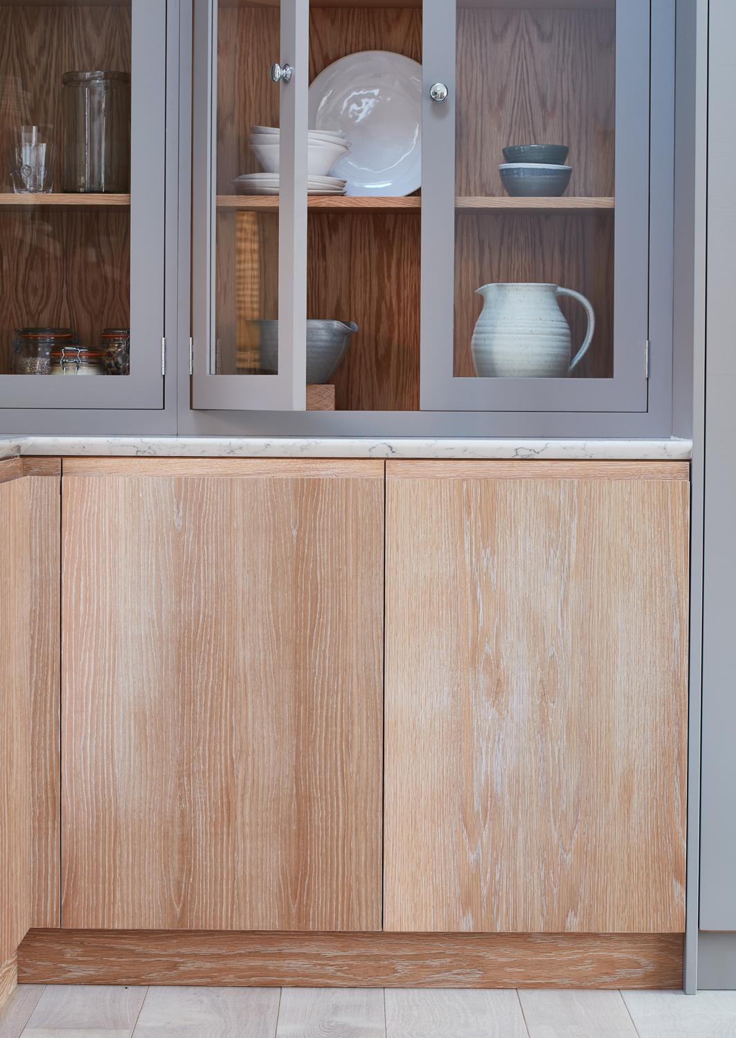 A kitchen setup featuring wooden lower cabinets with a light finish and a wooden display cabinet above with glass doors. The display cabinet houses white and blue dishes, including a jug, bowls, glasses, and plates. The countertop appears to be marble.