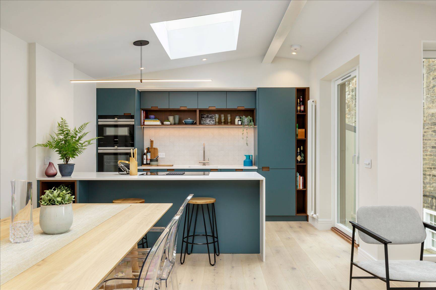 A modern kitchen and dining area featuring blue cabinets, a white countertop island with bar stools, a skylight, and wooden flooring. A dining table with transparent chairs and potted plants adds a touch of greenery to the space.