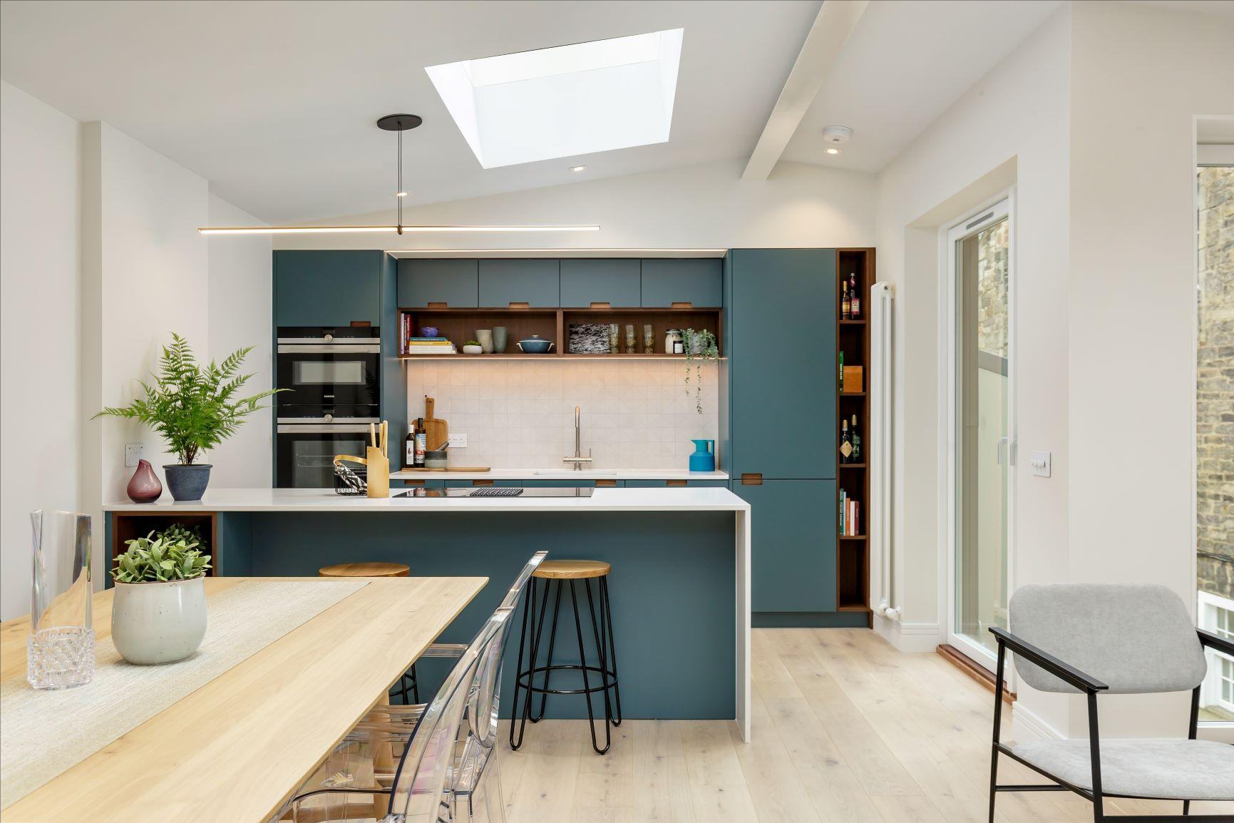 A modern kitchen and dining area featuring blue cabinets, a white countertop island with bar stools, a skylight, and wooden flooring. A dining table with transparent chairs and potted plants adds a touch of greenery to the space.