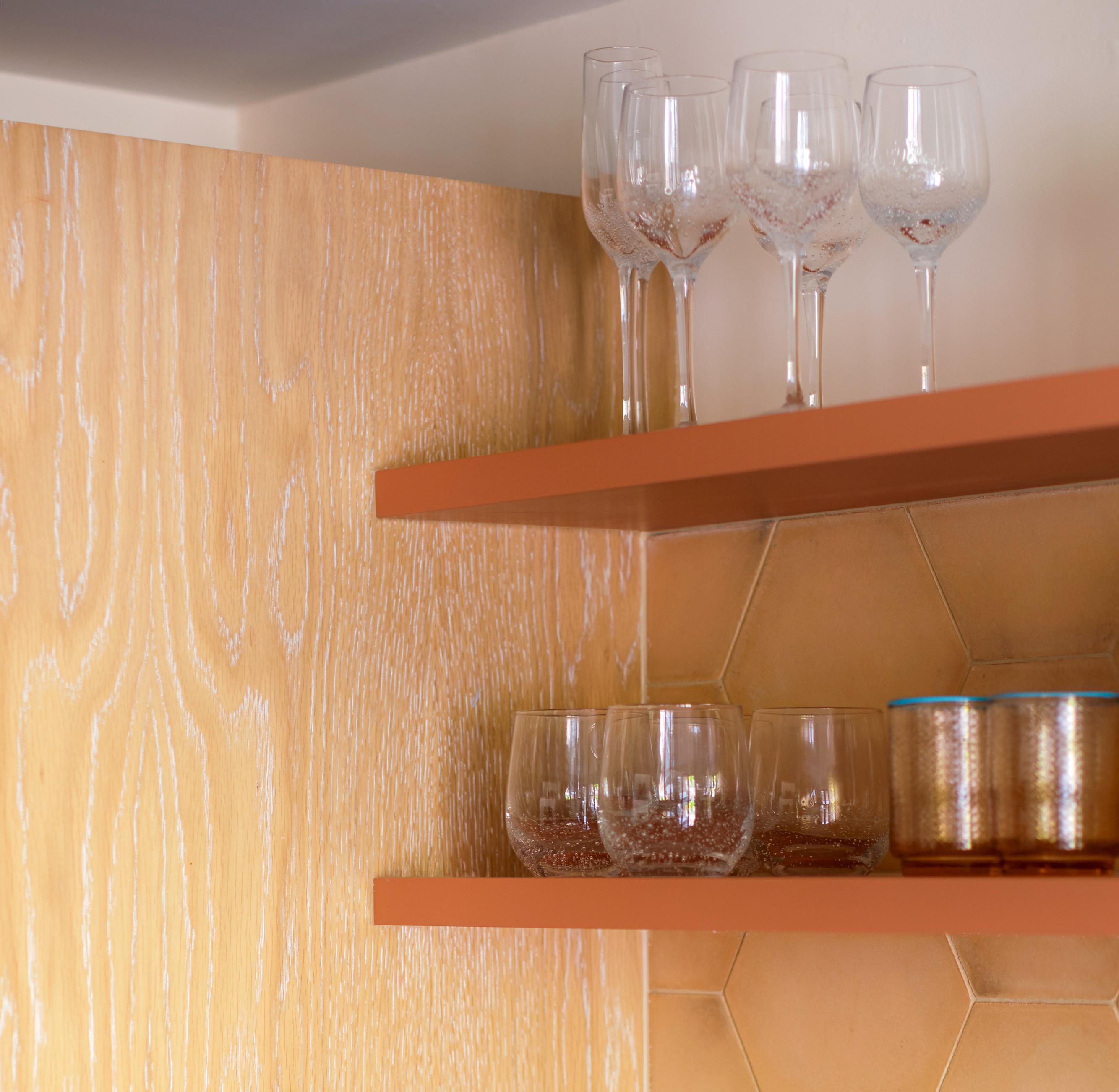 Two orange floating shelves hold clear wine and whiskey glasses, along with a metallic gold container, against a light wood and hexagonal tan tile wall.
