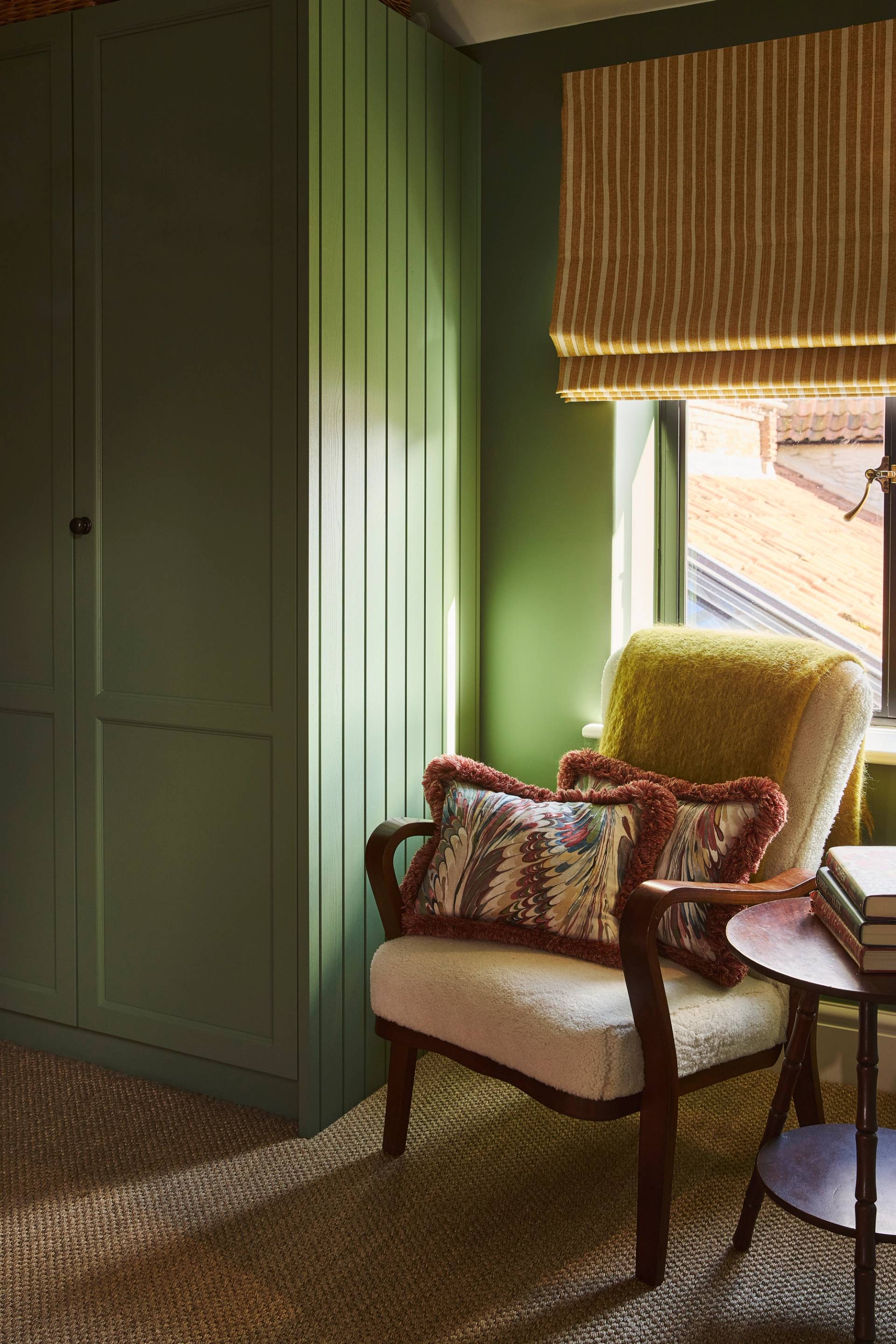 A cozy bedroom corner with olive green walls, a matching green wardrobe with baskets on top, a dark wood chest of drawers with a decorative mirror above, and a patterned armchair and carpet.