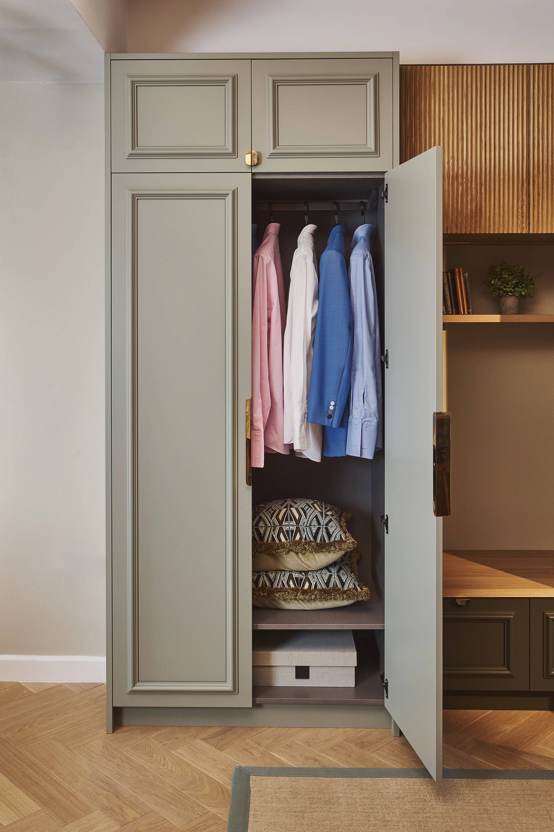 A wardrobe with open doors shows hanging shirts and jackets, a silver suitcase, patterned pillows, a few pairs of shoes, and storage boxes organized on shelves inside with one door closed