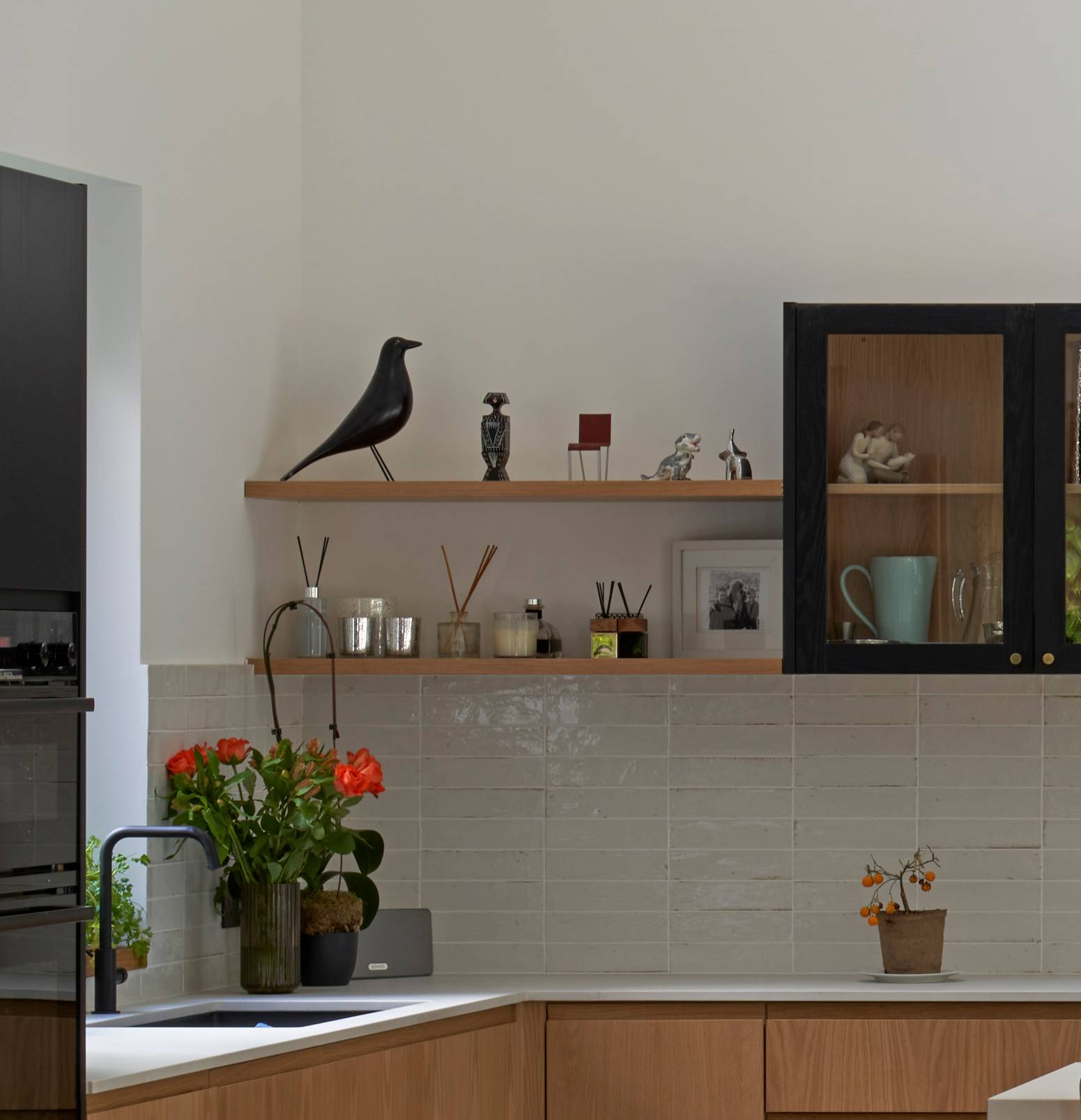 A modern kitchen with light wood cabinets, white countertops, and light gray tiled backsplash. A shelf holds a black bird sculpture, small items, and framed photos. Bright orange flowers and decorative items are placed on the counter.