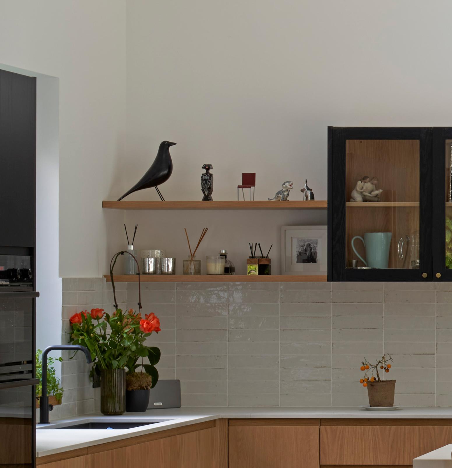A modern kitchen with light wood cabinets, white countertops, and light gray tiled backsplash. A shelf holds a black bird sculpture, small items, and framed photos. Bright orange flowers and decorative items are placed on the counter.