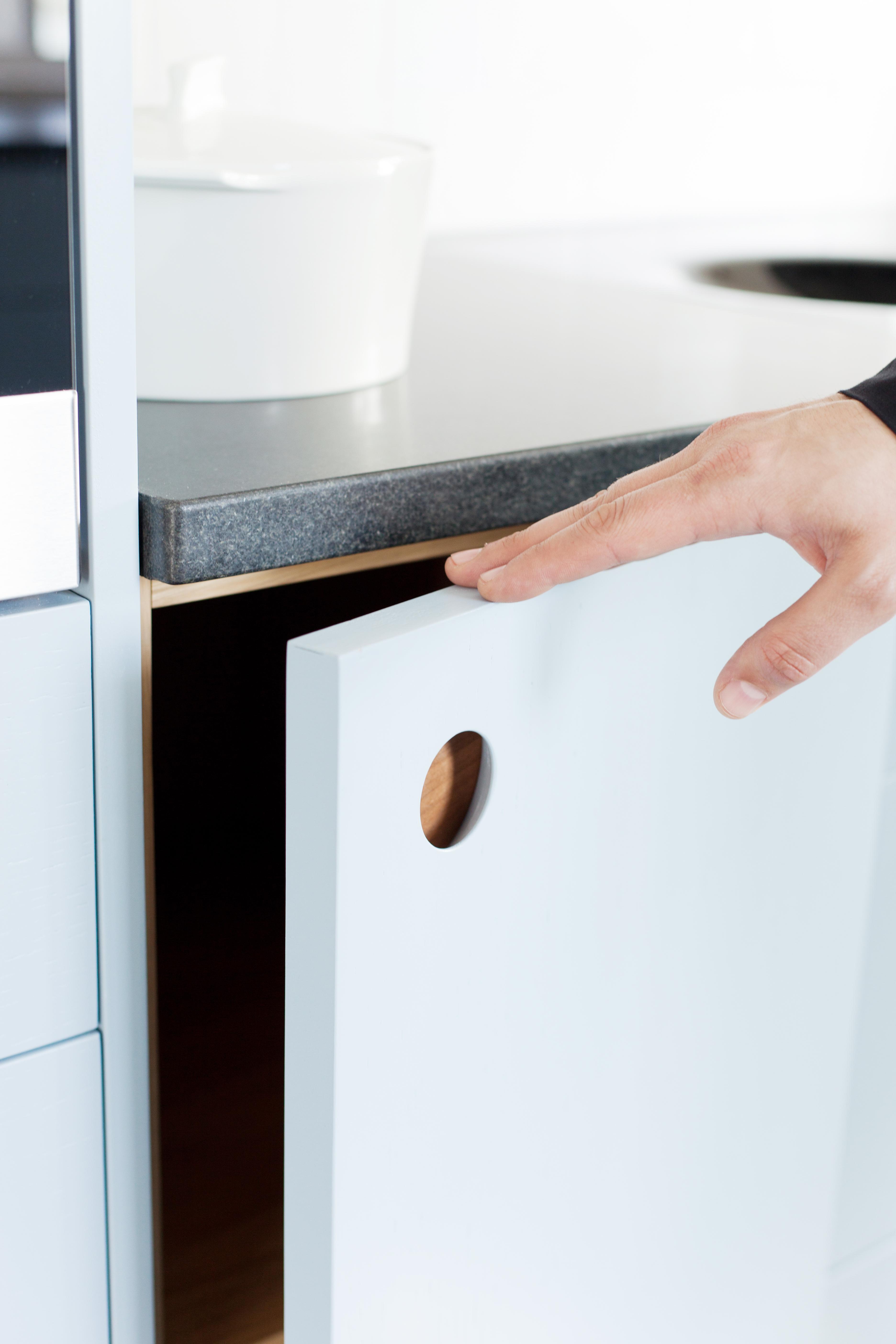 A close-up of a hand opening a light blue kitchen cabinet door, highlighting a circular cutout handle with a wood interior detail beneath a dark grey countertop.