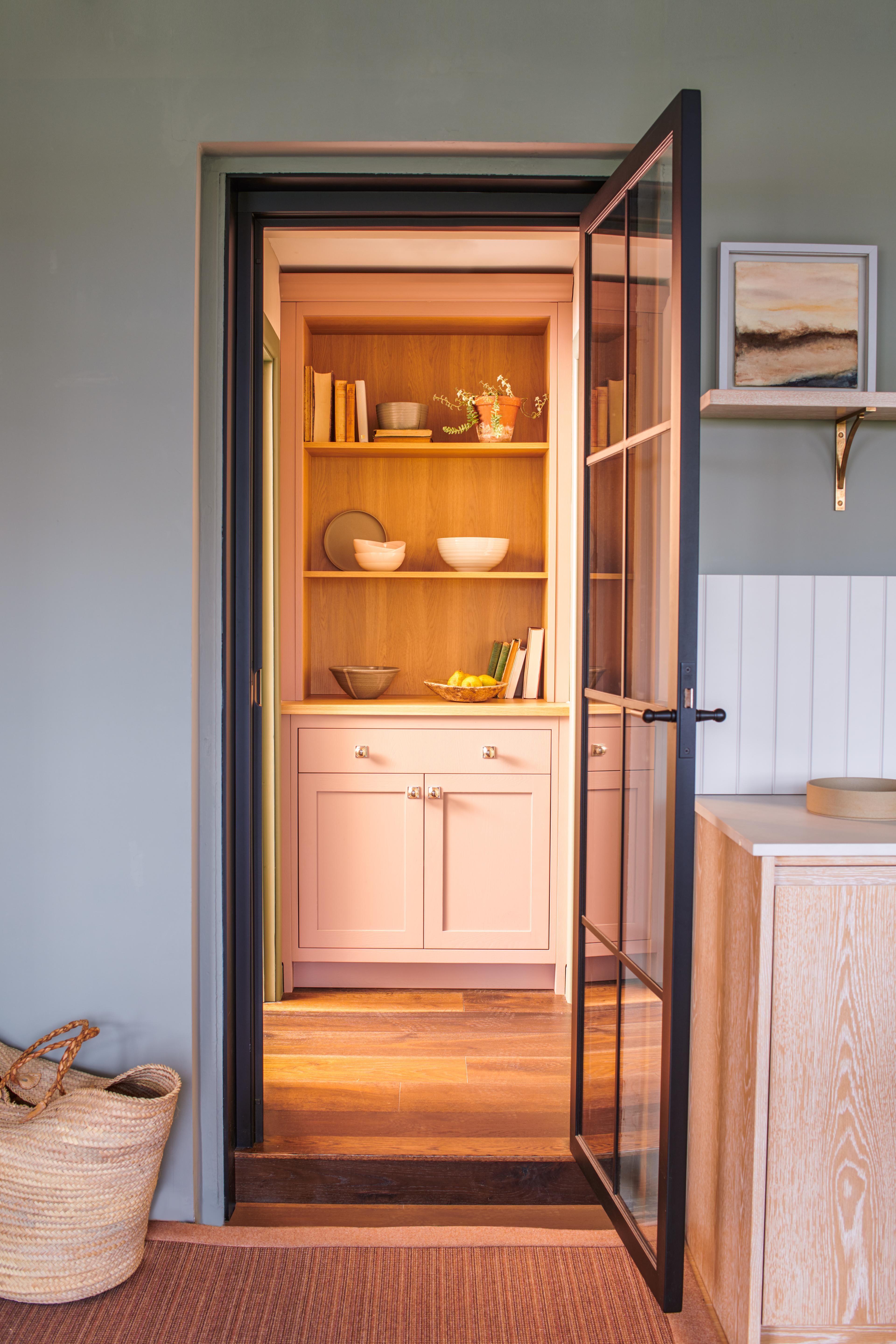 A glass door opens to a cozy nook with light pink cabinets and wooden shelves holding books and decorative items. Soft lighting brightens the space, which is framed by green walls and surrounded by woven baskets and light wood accents.