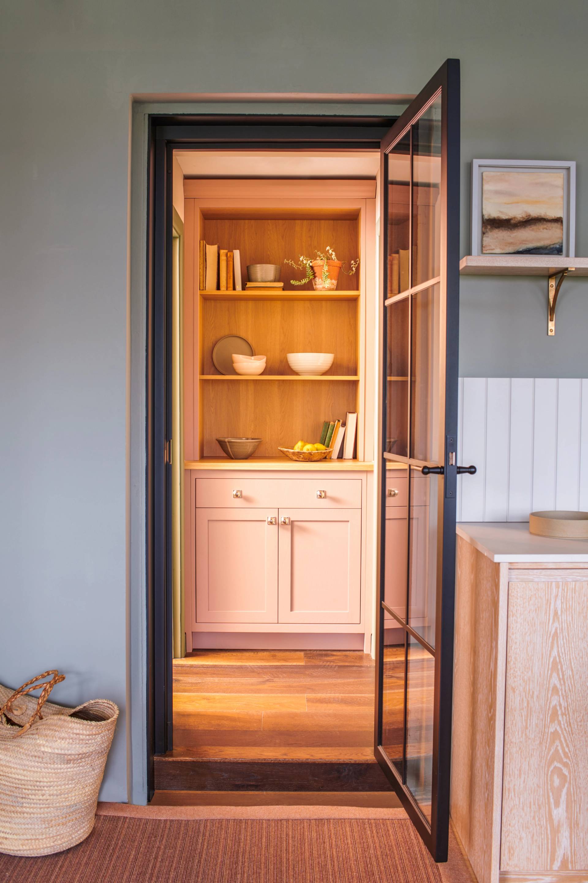 A glass door opens to a cozy nook with light pink cabinets and wooden shelves holding books and decorative items. Soft lighting brightens the space, which is framed by green walls and surrounded by woven baskets and light wood accents.