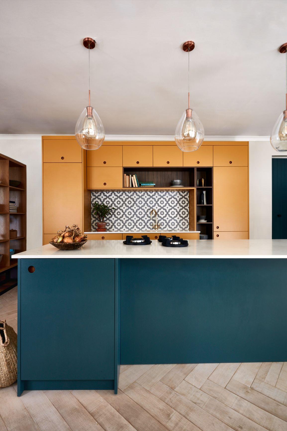 Modern kitchen with a blue island and white countertop. The background features mustard-colored cabinets and a patterned backsplash. Three pendant lights hang above the island, and a fruit bowl is placed on the counter. Light wood flooring completes the look.