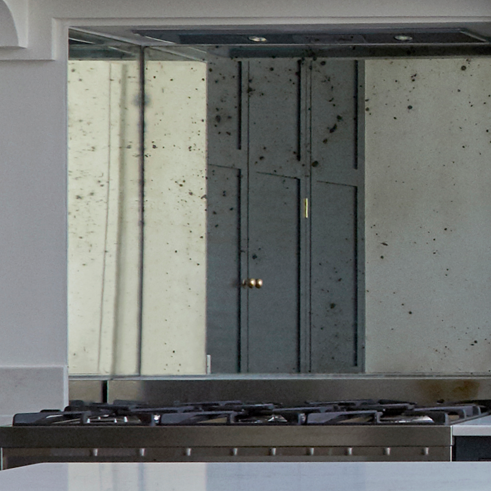 A close-up of a kitchen stove with a tarnished, speckled mirror backsplash reflecting dark cabinetry and a brass doorknob. The countertop in the foreground is white.