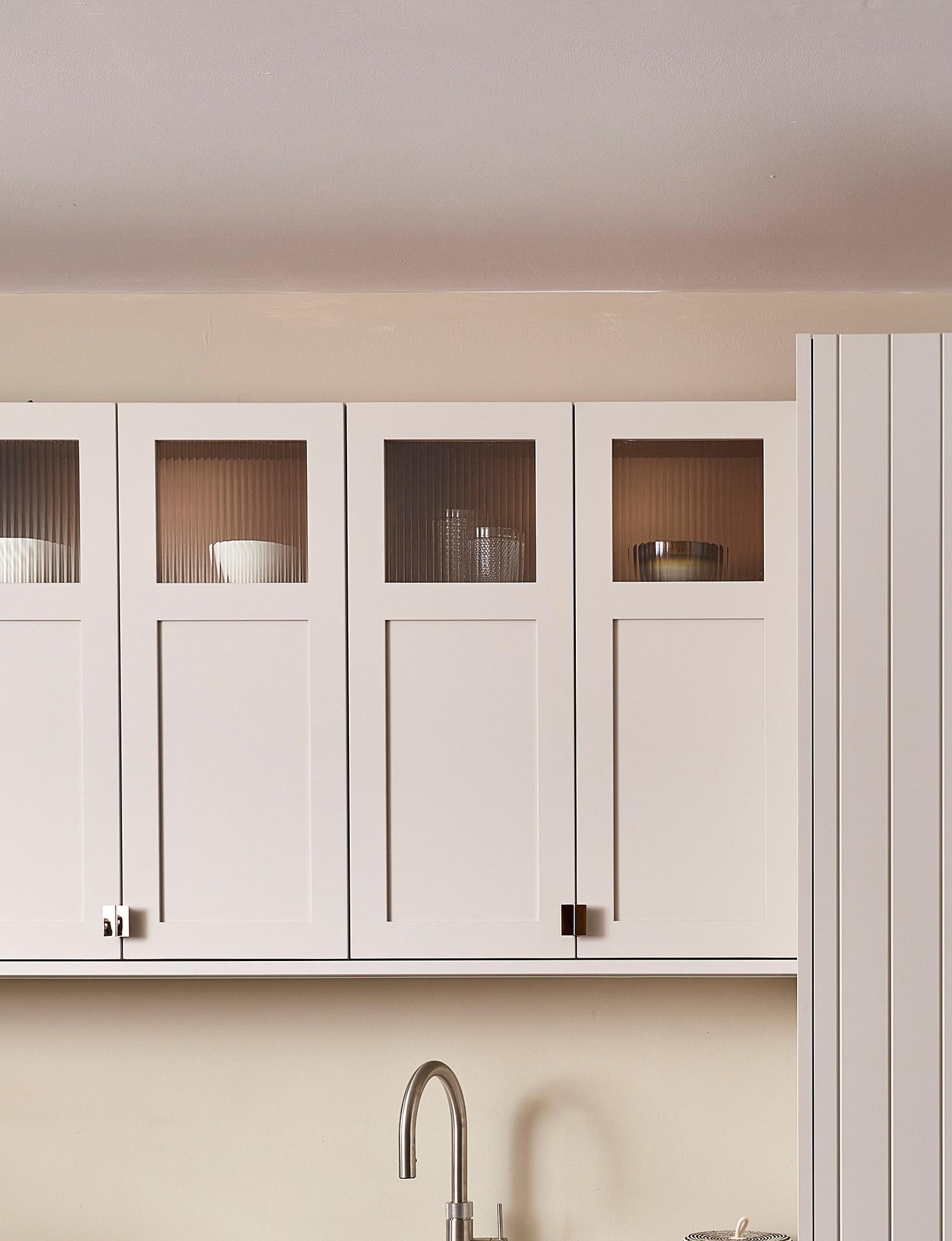 White kitchen cabinets with frosted glass panels at the top, displaying white bowls and a small bowl inside. Below the cabinets is a modern stainless steel faucet and sink, with a cream-colored backsplash.