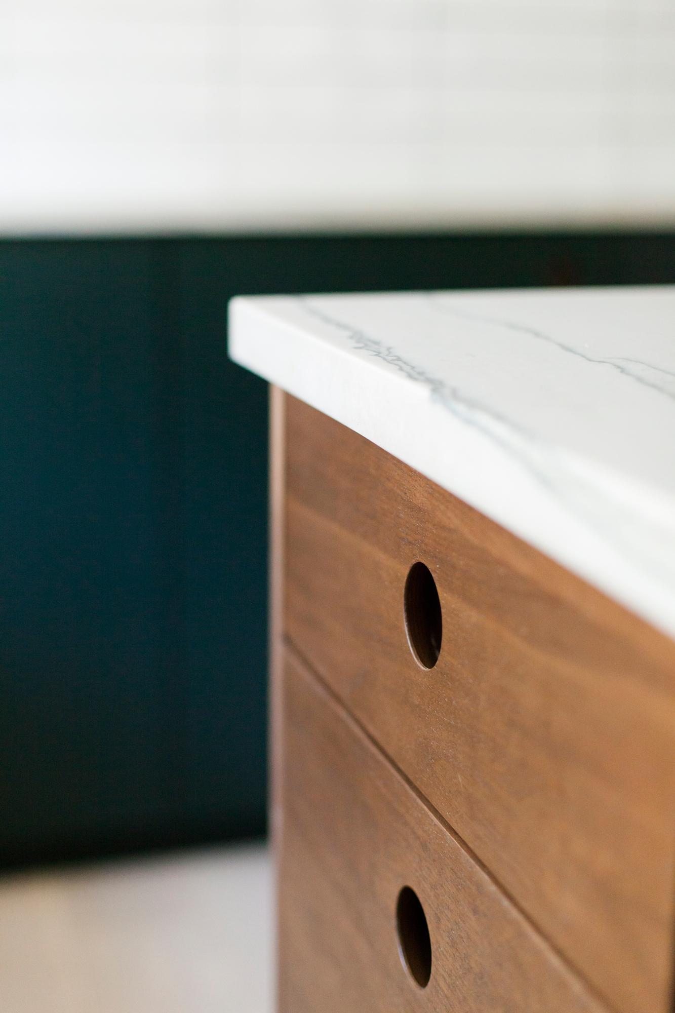 Close-up of a wooden cabinet with round cutout handles and a white marble countertop, set against a blurred dark background.