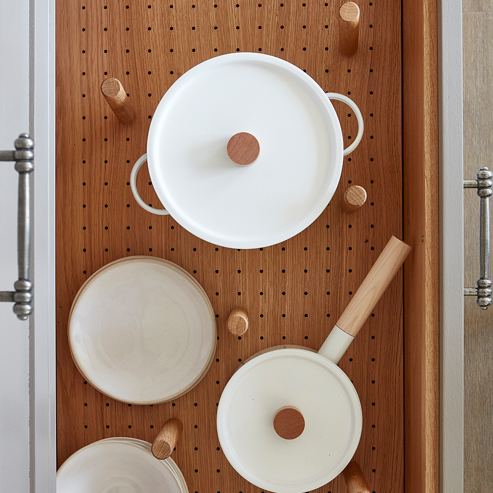 A kitchen drawer with a pegboard organizer holding white ceramic pots and pans with wooden handles and lids, arranged neatly with wooden pegs.