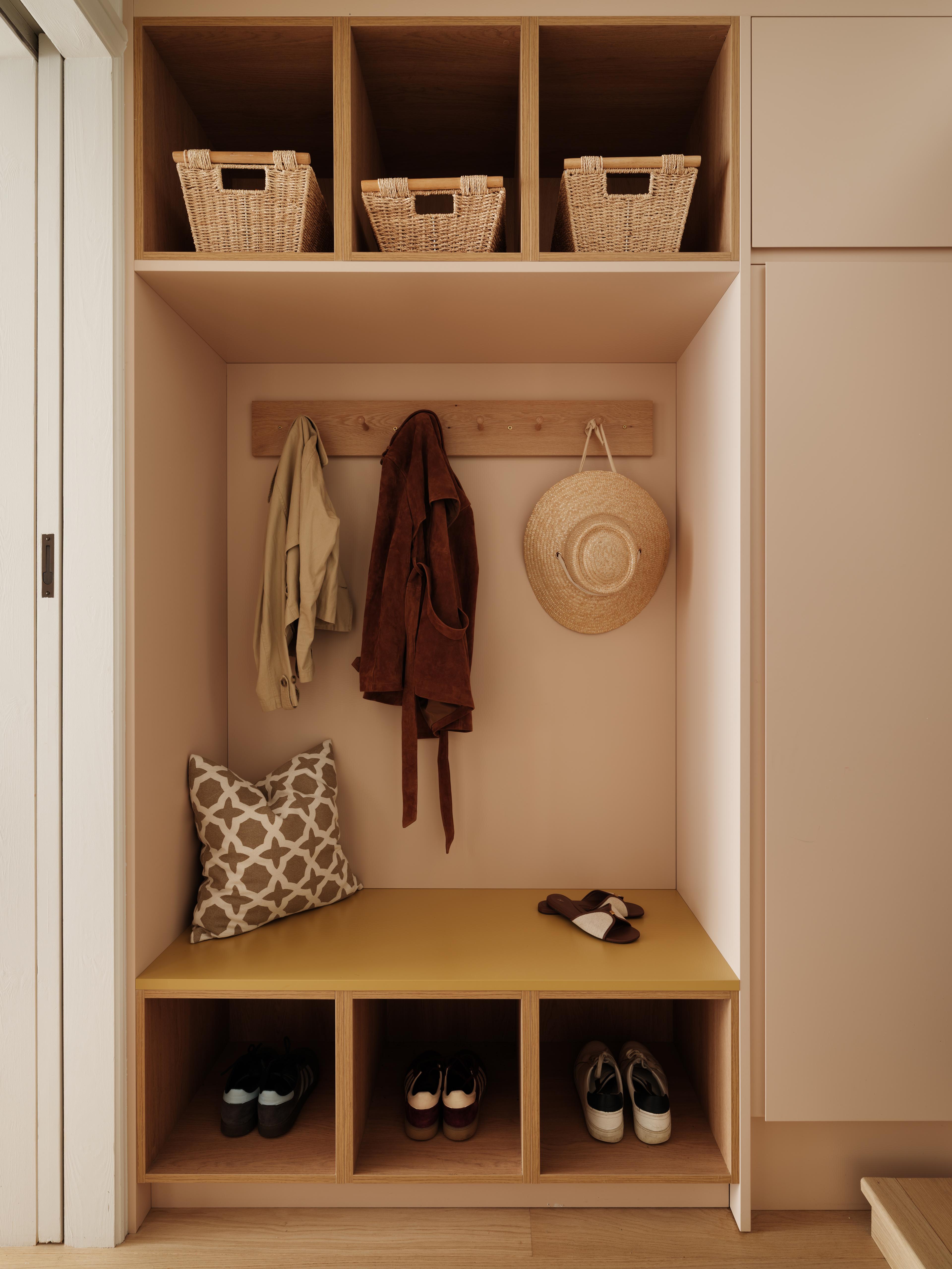 A stylish mudroom with gray cabinets, a drawer slightly open, and a shelf displaying a hat and a figurative painting. A pair of boots and a polo mallet are on the wooden floor near a beige rug. Natural light streams through windows above.