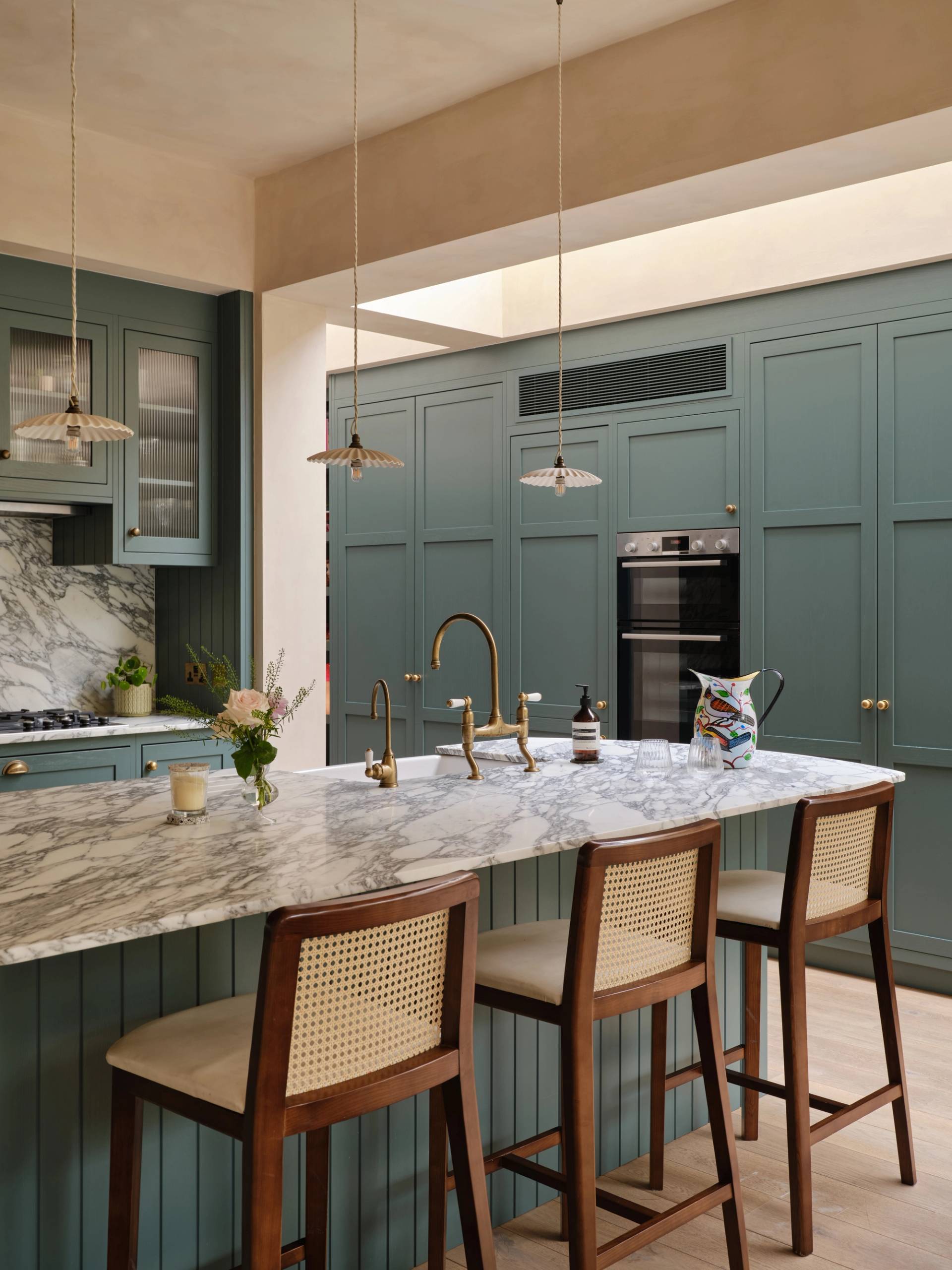 A cozy kitchen with a mix of styles. Light blue cabinets complement a marbled countertop and backsplash. A wooden shelf holds books and dishes, while a maroon island features a cutting board and red pot. Plants hang above the sink for decoration.
