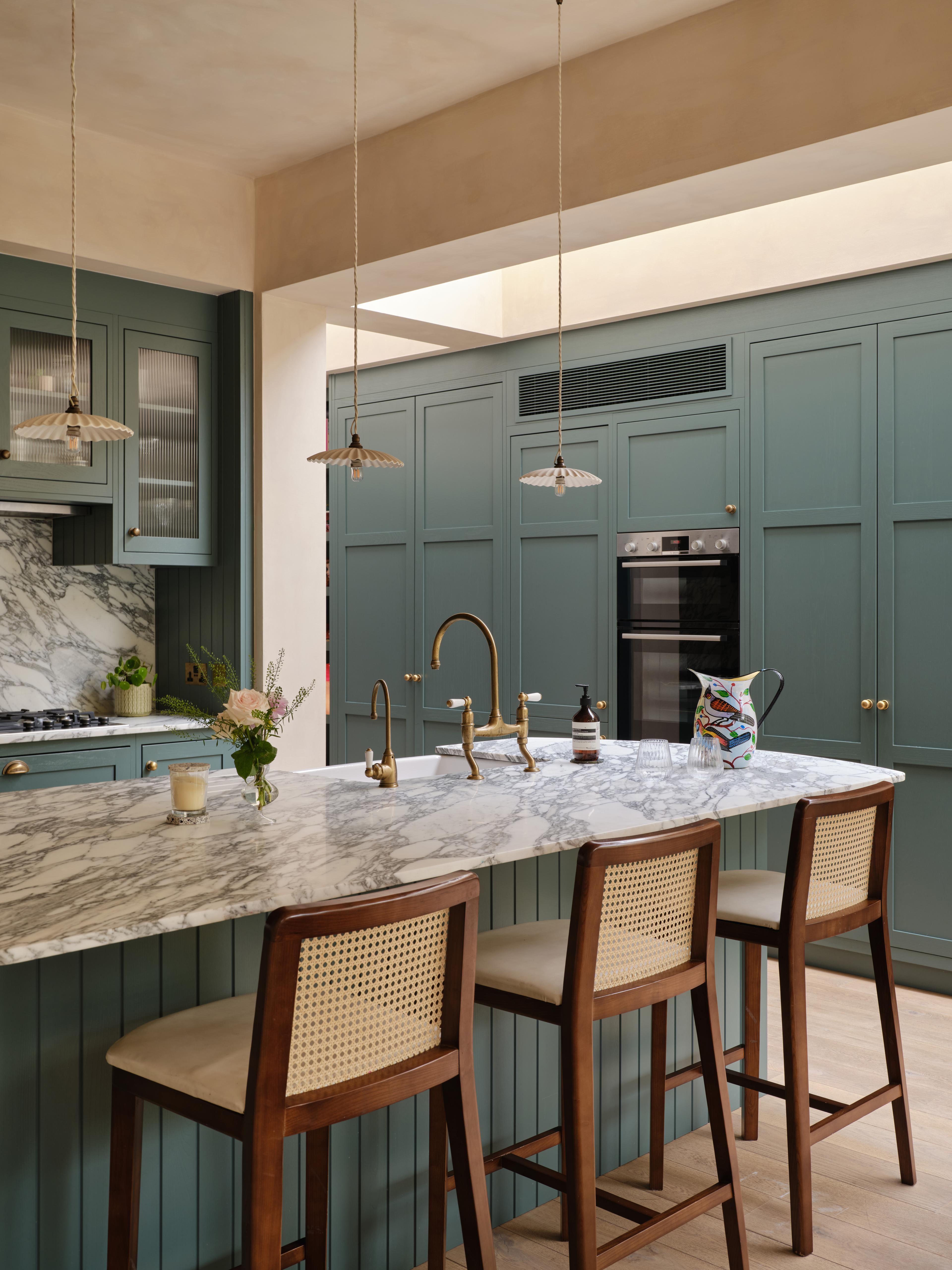 A cozy kitchen with a mix of styles. Light blue cabinets complement a marbled countertop and backsplash. A wooden shelf holds books and dishes, while a maroon island features a cutting board and red pot. Plants hang above the sink for decoration.