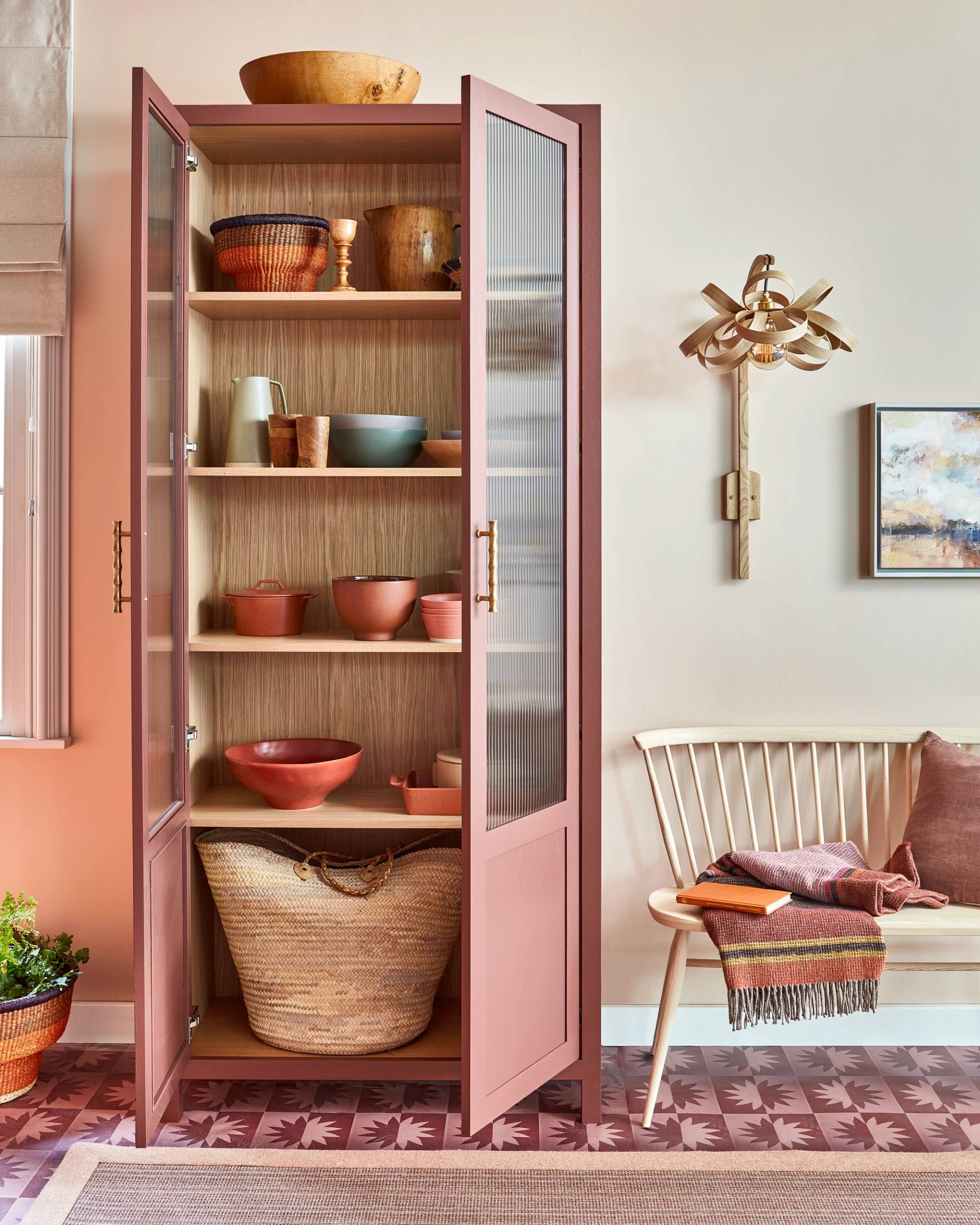 A tall wooden cabinet with pink doors stands open, revealing neatly arranged bowls and baskets. Beside it is a wooden chair with a folded blanket. A unique wall sconce and a small painting decorate the beige wall. The floor has a patterned design