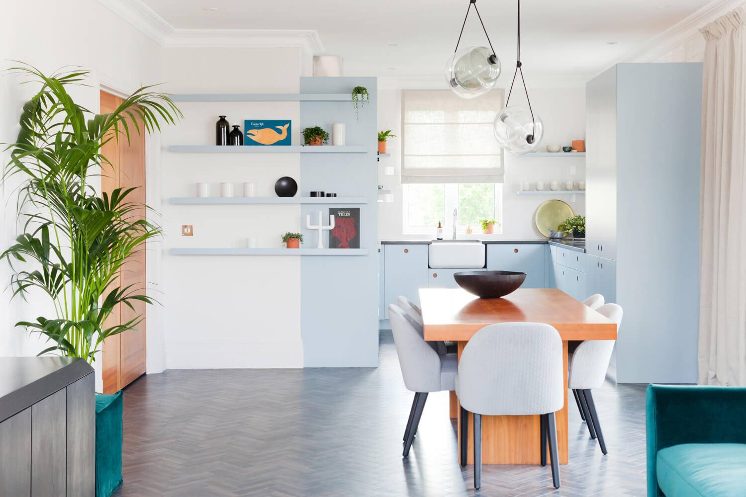 Modern kitchen with pastel blue cabinets and open shelving displaying decor items. A wooden dining table with gray chairs is centered under unique pendant lights. A large potted plant decorates the space, with a window providing natural light in the background.