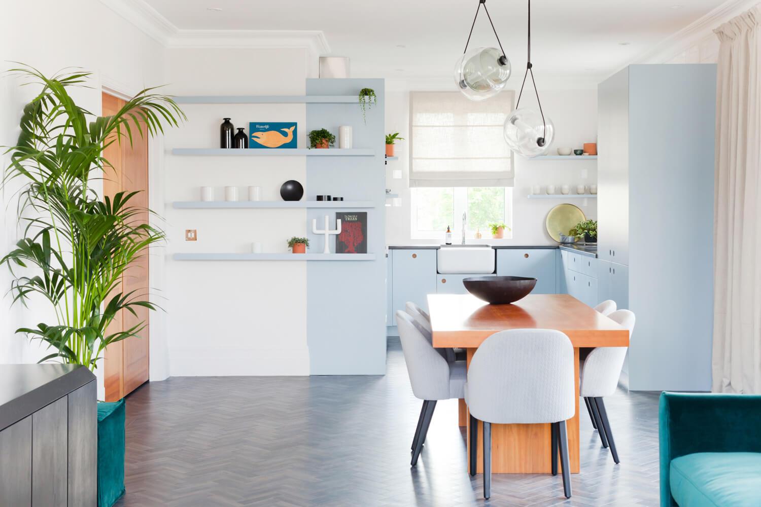Modern kitchen with pastel blue cabinets and open shelving displaying decor items. A wooden dining table with gray chairs is centered under unique pendant lights. A large potted plant decorates the space, with a window providing natural light in the background.