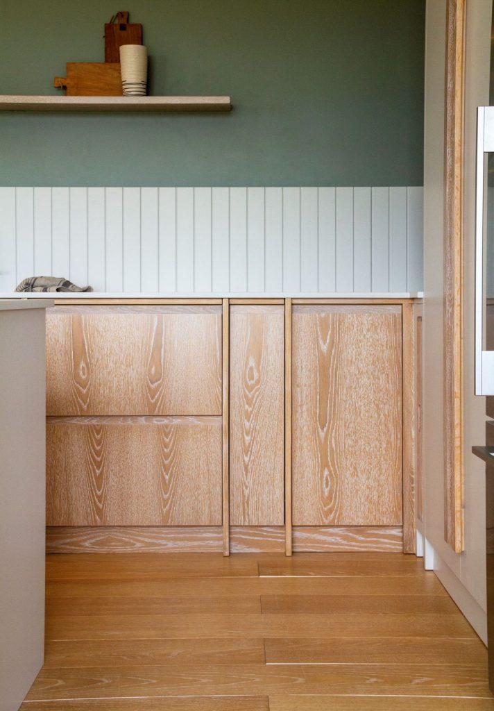 Light wood kitchen cabinets featuring prominent grain patterns, set below white vertical tongue-and-groove paneling and a sage green wall with an open floating shelf.