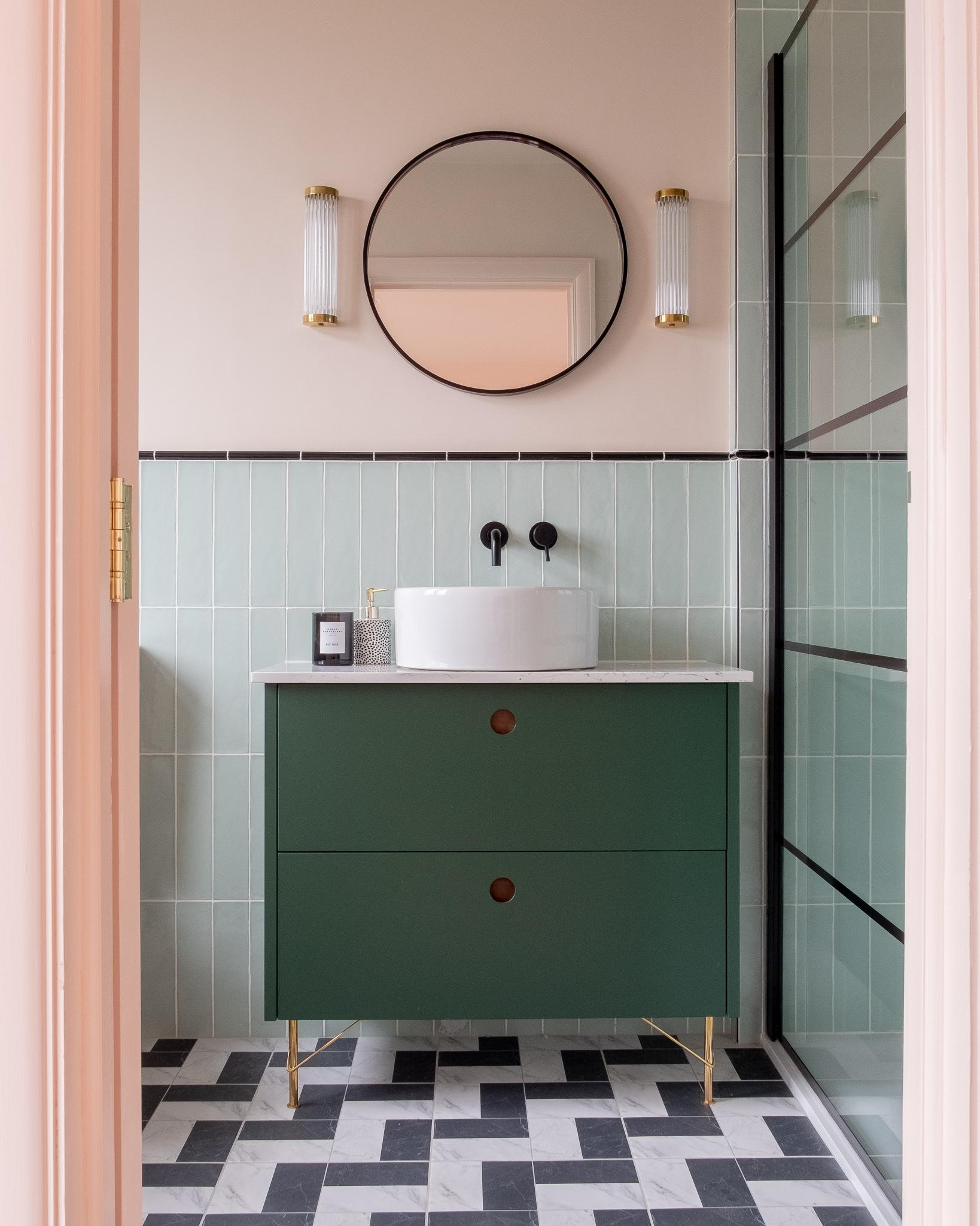 Modern bathroom featuring a dark green vanity with a round white sink. Two cylindrical wall lights flank a round mirror above. Light green tiled walls and black-and-white checkered floor tiles complete the design.