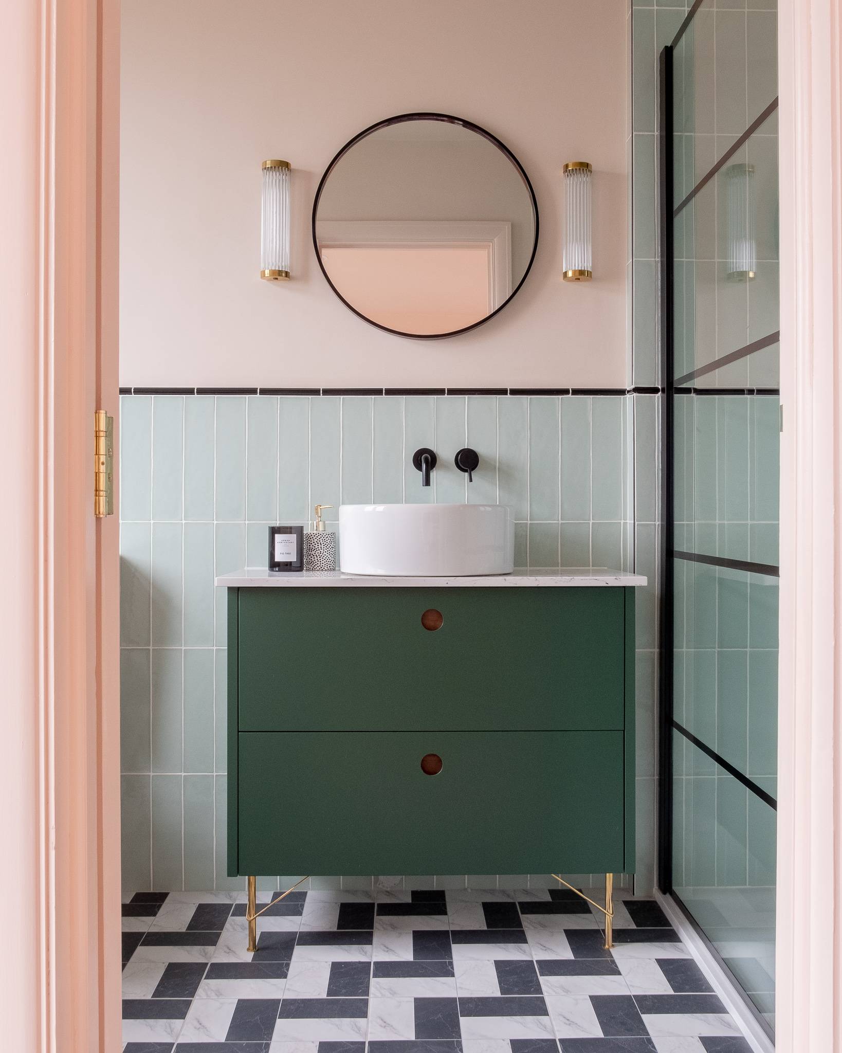 Modern bathroom featuring a dark green vanity with a round white sink. Two cylindrical wall lights flank a round mirror above. Light green tiled walls and black-and-white checkered floor tiles complete the design.