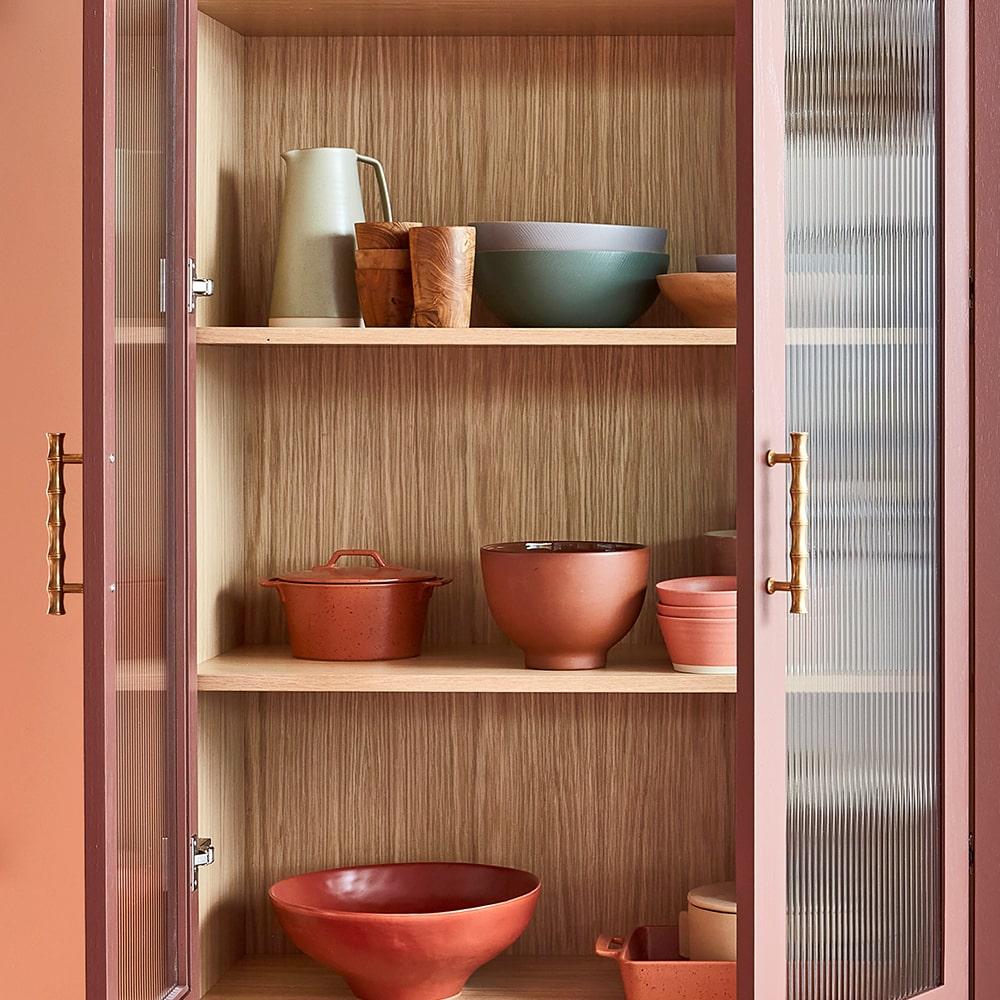 A wooden kitchen cabinet with ribbed glass doors displays neatly arranged ceramic bowls, plates, a pitcher, and a wooden cup in earthy tones on three shelves.